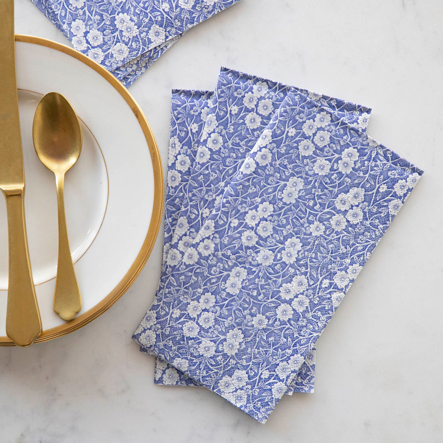 A stack of three Blue Calico Guest Napkins fanned out next to a gold-rimmed plate with gold flatware. 