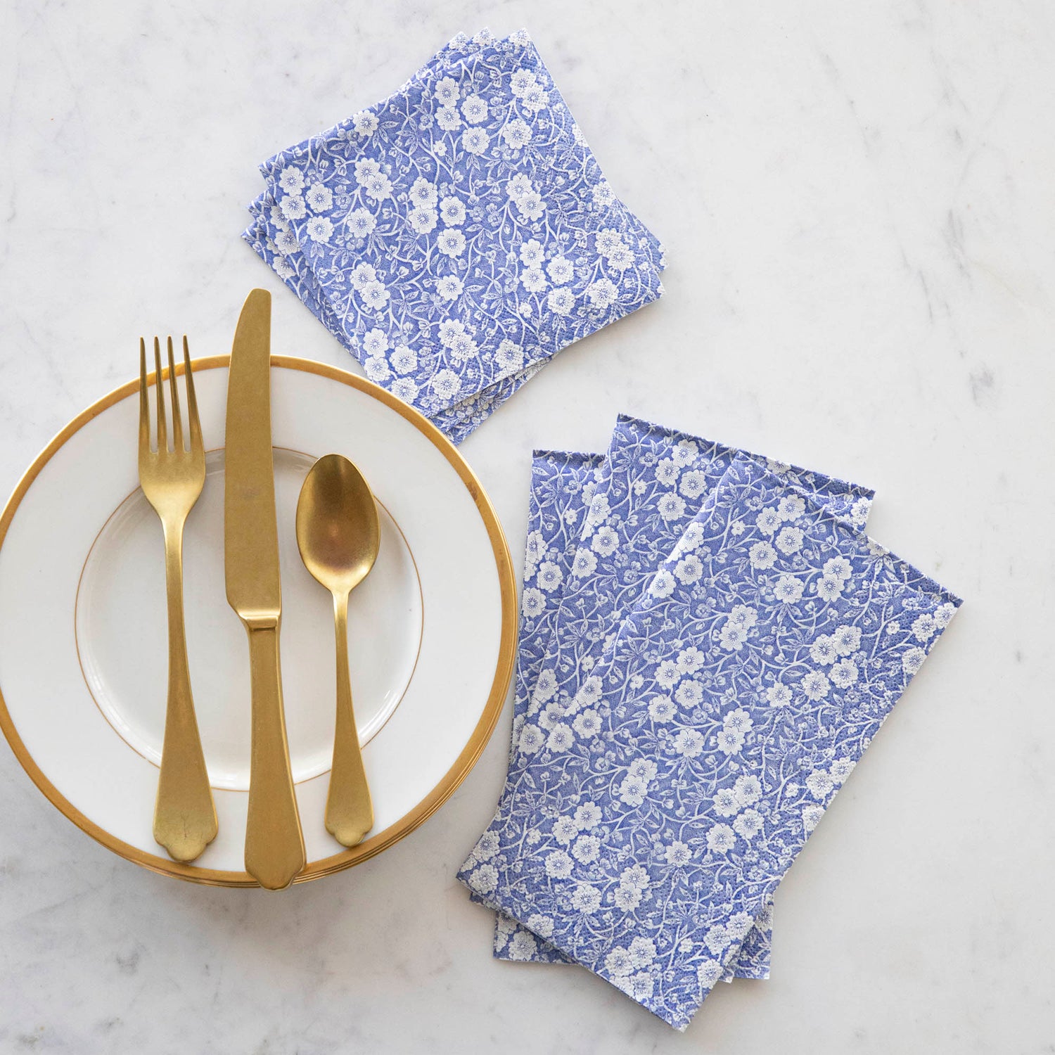 A stack of three Blue Calico Guest Napkins and a stack of three Blue Calico Cocktail Napkins fanned out next to a gold-rimmed plate with gold flatware.