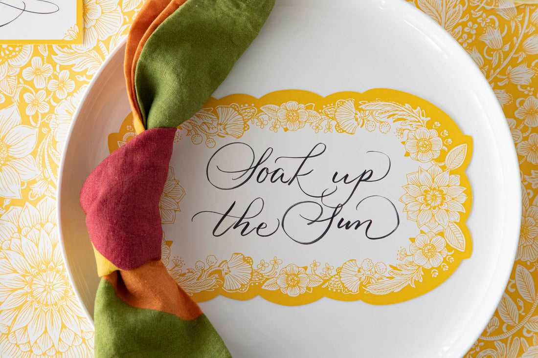 Close-up of a Spring Blooms Table Card with &quot;Soak up the sun&quot; written on it in lovely script resting on the plate of a vibrant place setting.