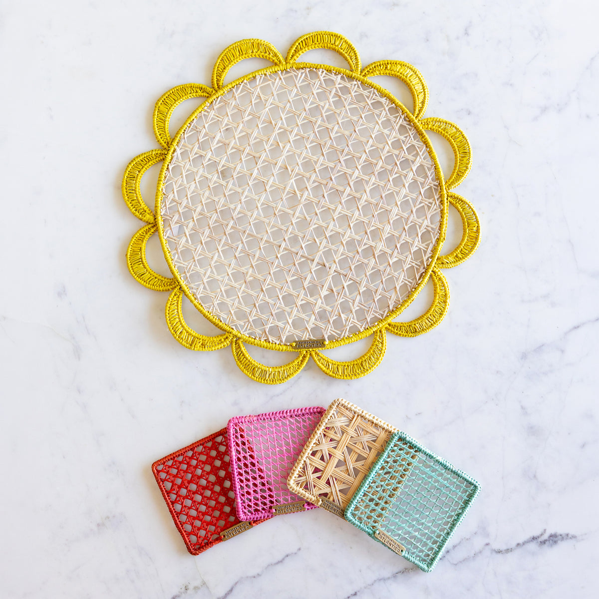 A round yellow woven placemat with a scalloped gold border is displayed on a white marble surface, with four smaller matching coasters in red, pink, tan, and mint arranged below it.