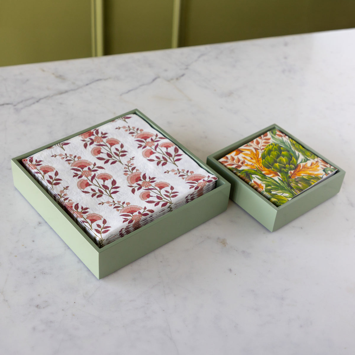 Two square gift boxes with pale sage green lids sit on a white marble countertop. One box contains folded napkins with a rust and brown floral pattern on white, and the other contains folded paper towels or cloths with a green, yellow, and orange botanical print.
