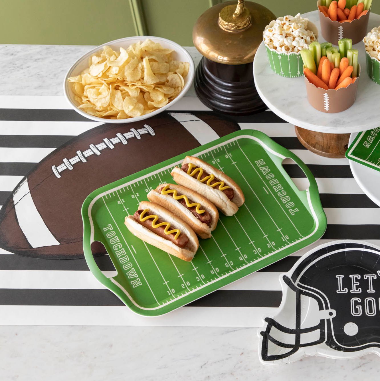 Football field tray of hotdogs on top of a black stripe runner and die-cut football placemat, surrounded by various snacks.