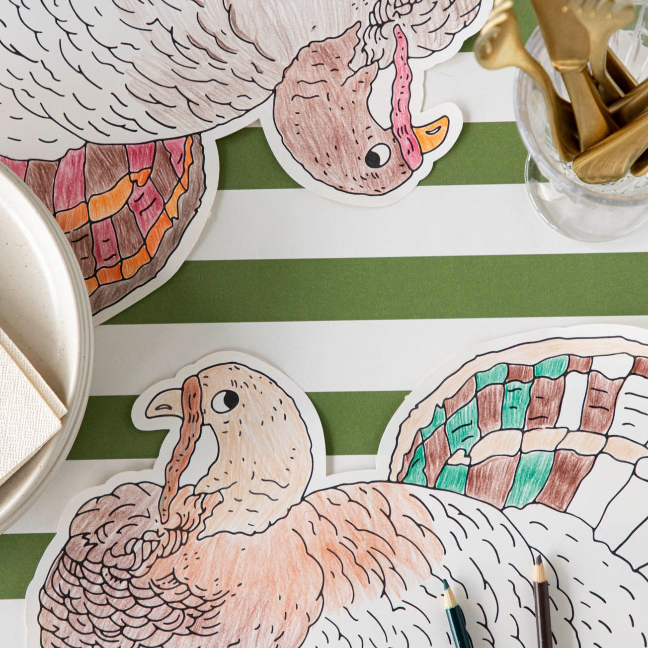 Die-cut coloring turkey placemat partially colored in with 2 colored pencils laying atop it on a green stripe runner.