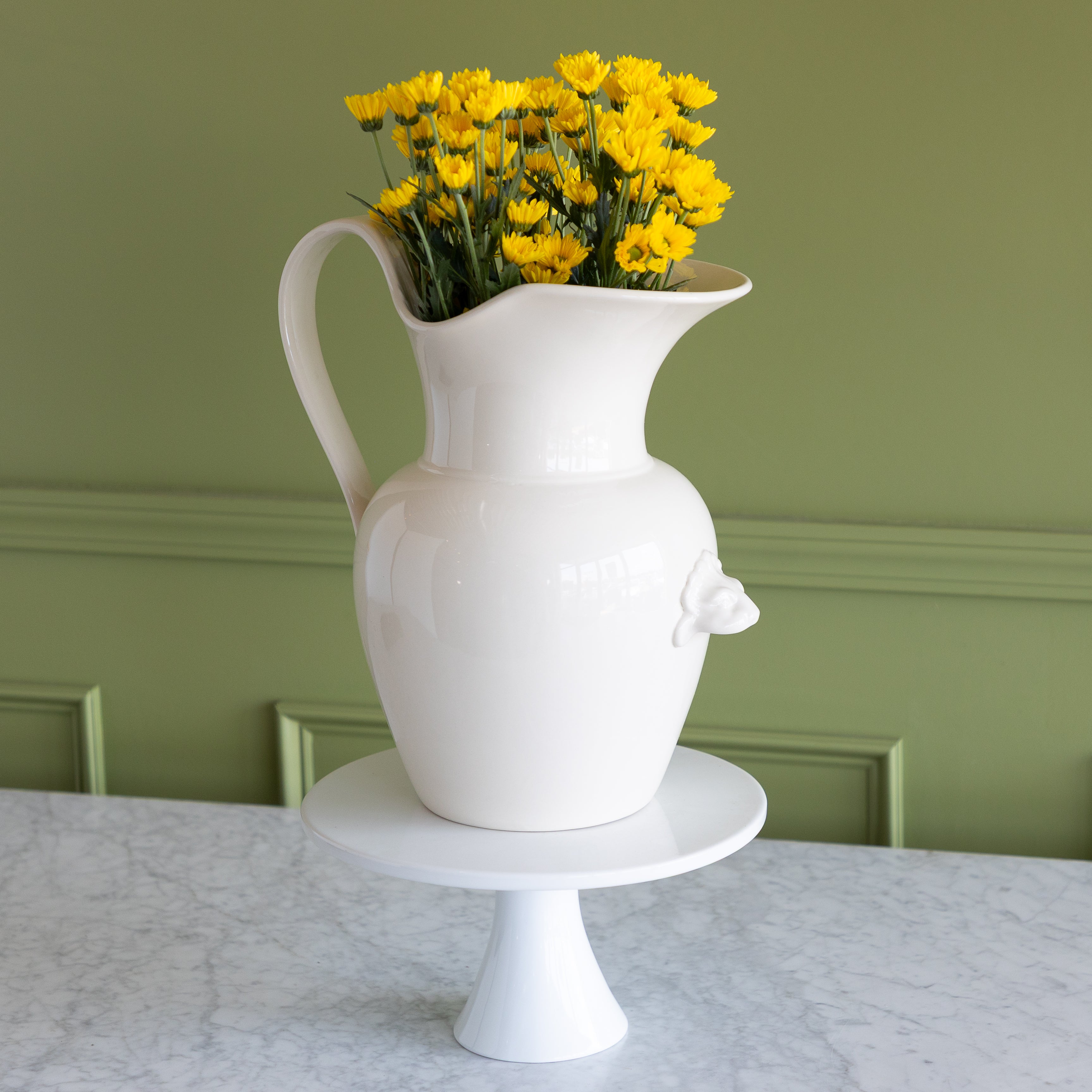 White pitcher with yellow flowers on a marble surface against a green wall.
