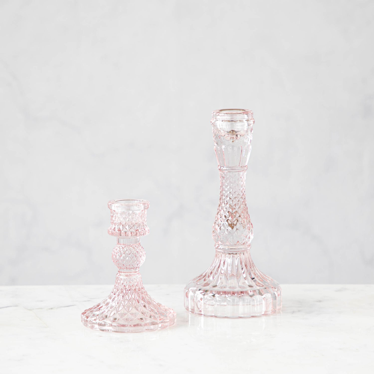 Two pink glass candlesticks with a geometric pattern, one short and one tall, on a marble surface.