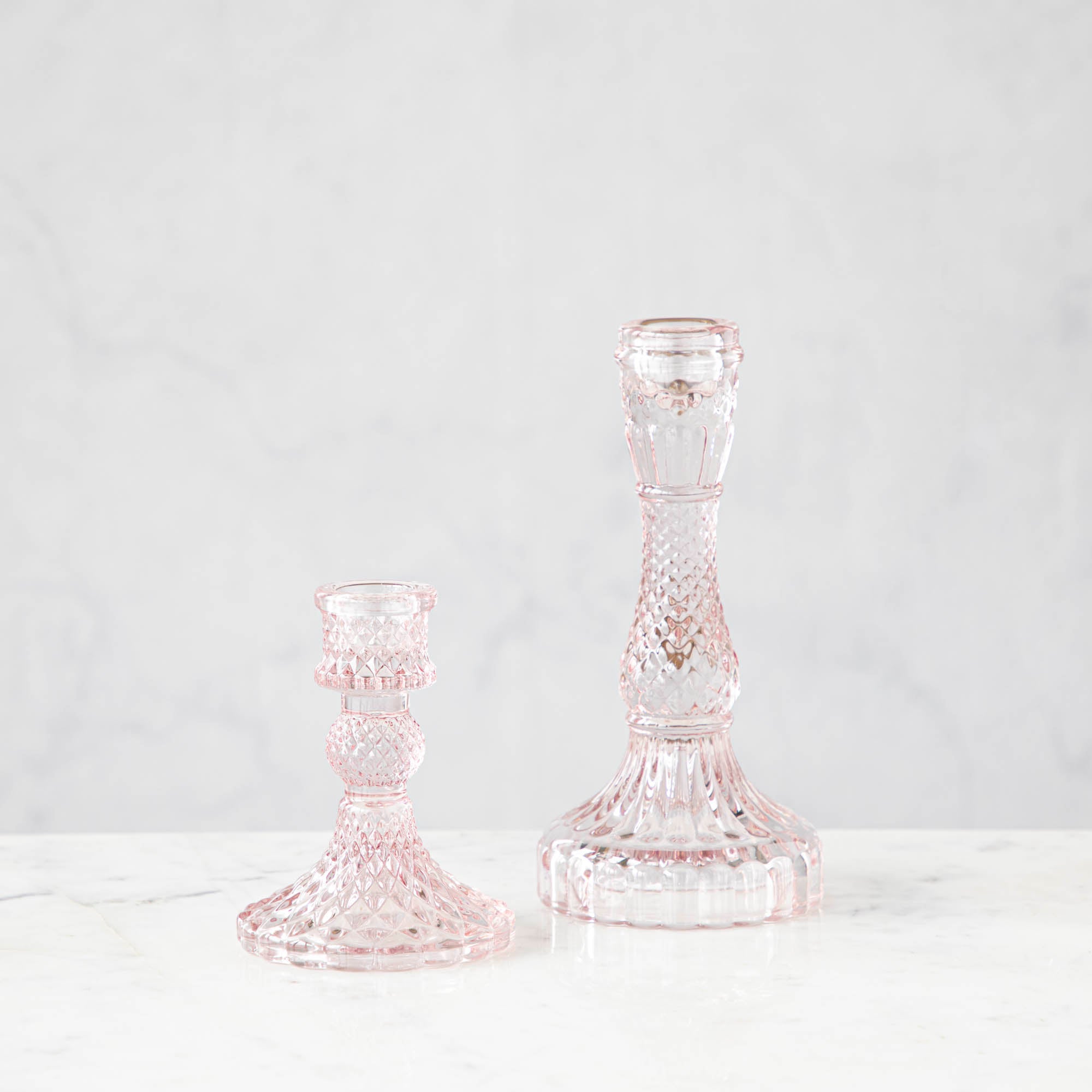 Two pink glass candlesticks with a geometric pattern, one short and one tall, on a marble surface.