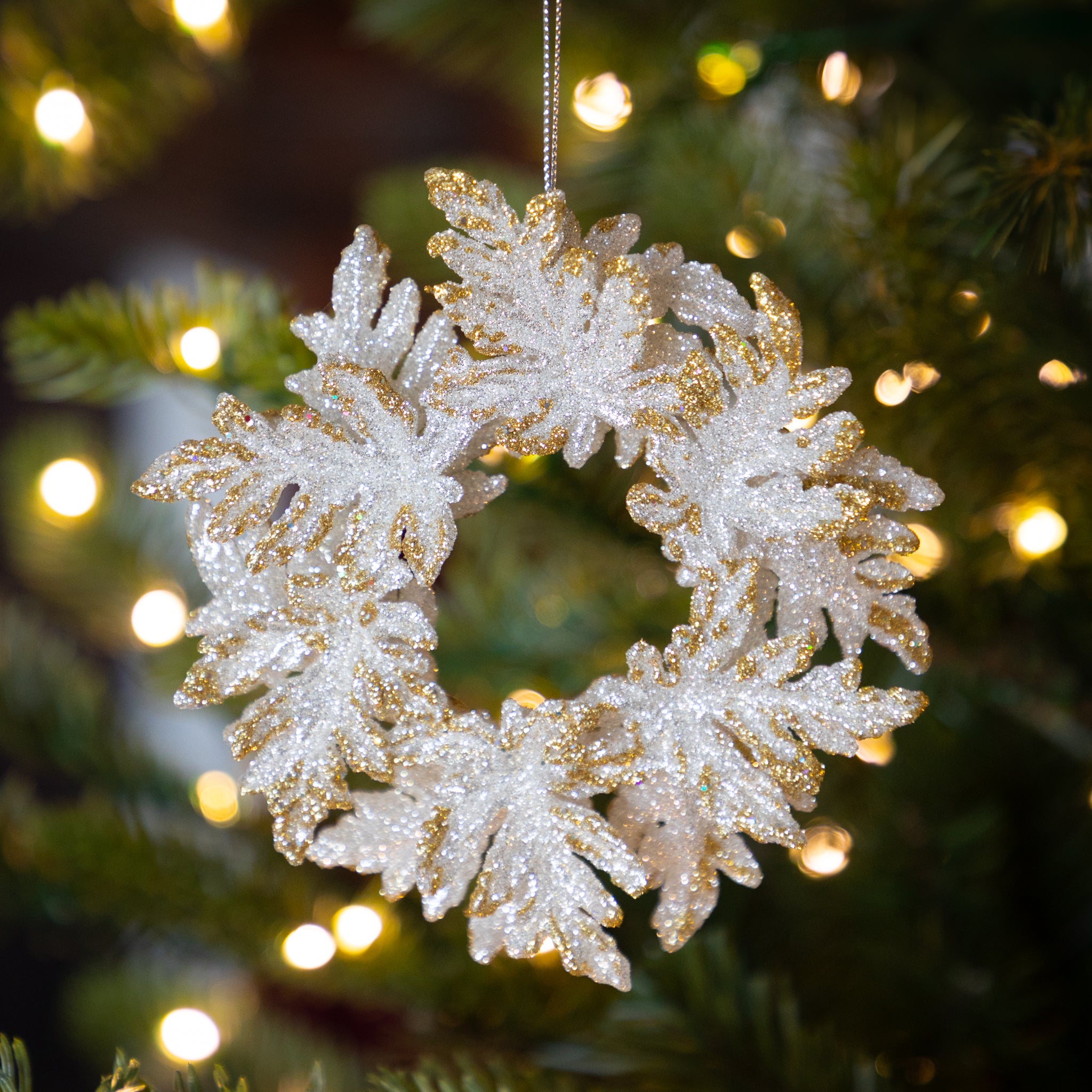 Decorative silver wreath with gold tips ornament on a Christmas tree with blurred lights in the background.