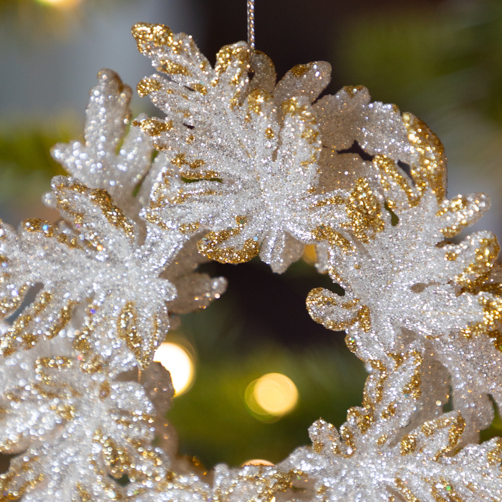 Decorative silver wreath with gold tips ornament on a Christmas tree with blurred lights in the background.