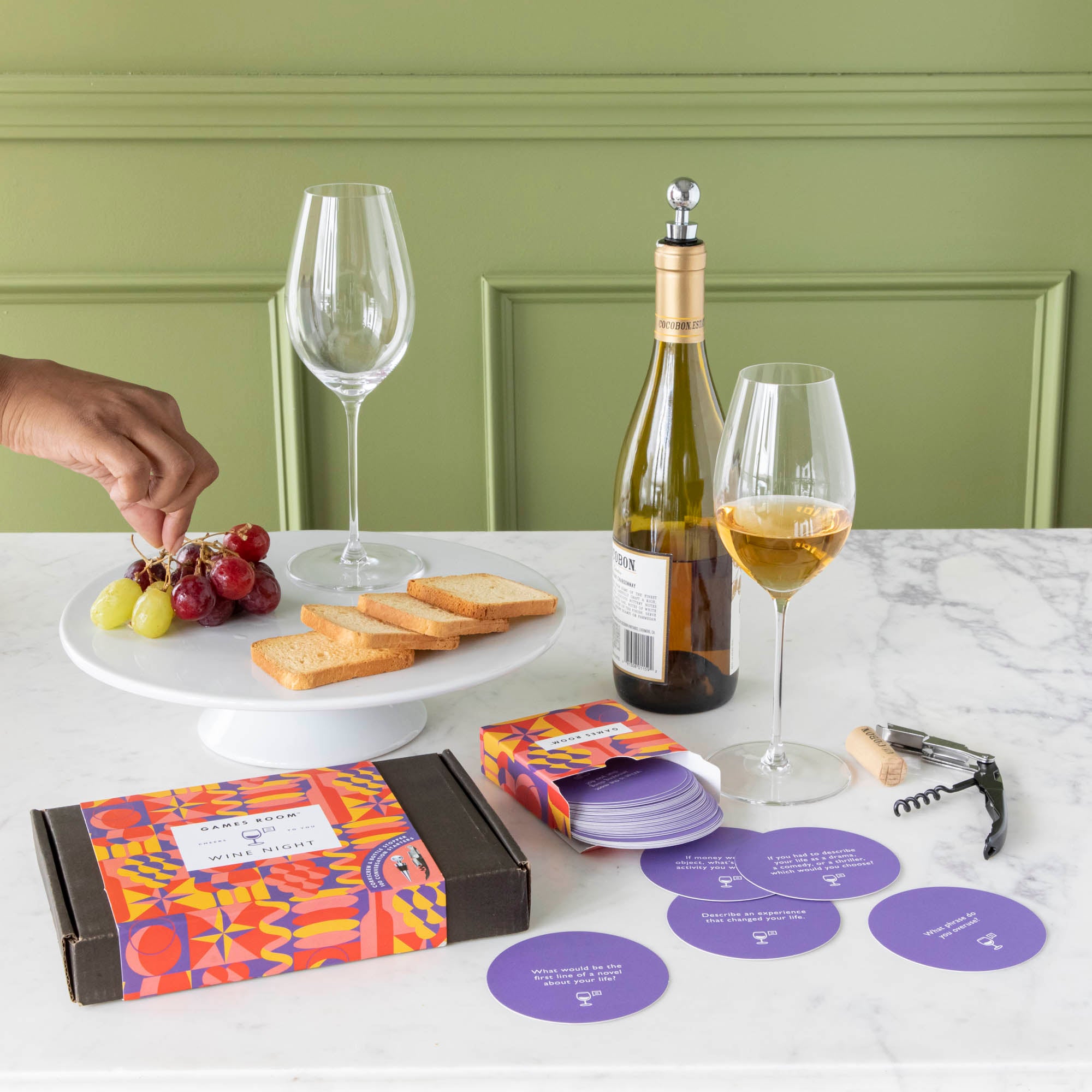 A gift set for wine nights displayed on a table, including a corkscrew, a bottle stopper, a glass of wine, a bottle of wine, bread, and grapes. The set comes in a colorful box with conversation starter cards visible.
