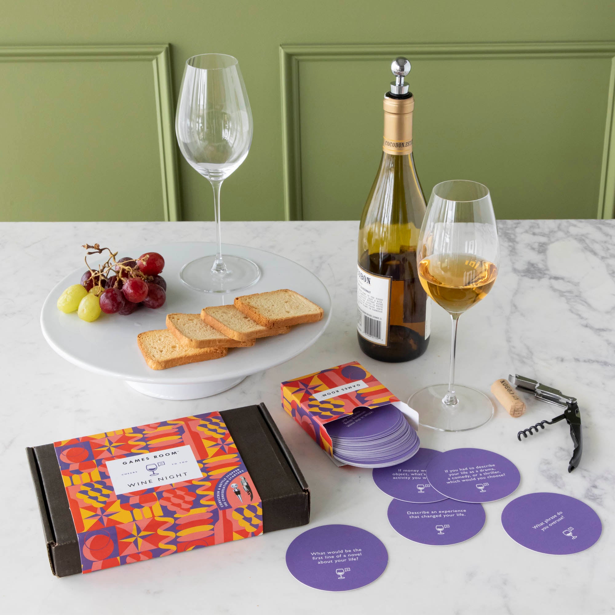 A gift set for wine nights displayed on a table, including a corkscrew, a bottle stopper, a glass of wine, a bottle of wine, bread, and grapes. The set comes in a colorful box with conversation starter cards visible.