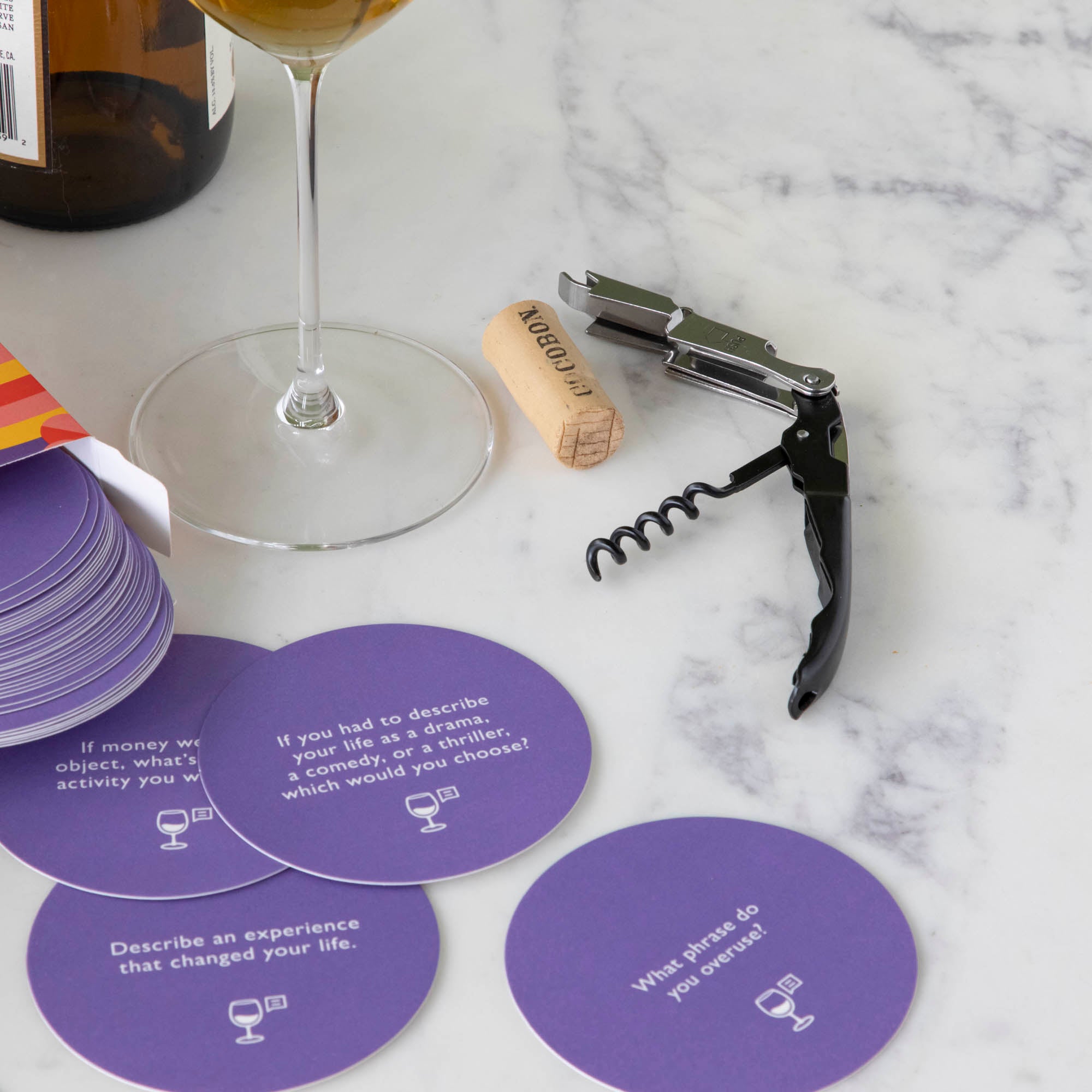 A gift set for wine nights displayed on a table, including a corkscrew, a bottle stopper, a glass of wine, a bottle of wine, bread, and grapes. The set comes in a colorful box with conversation starter cards visible.