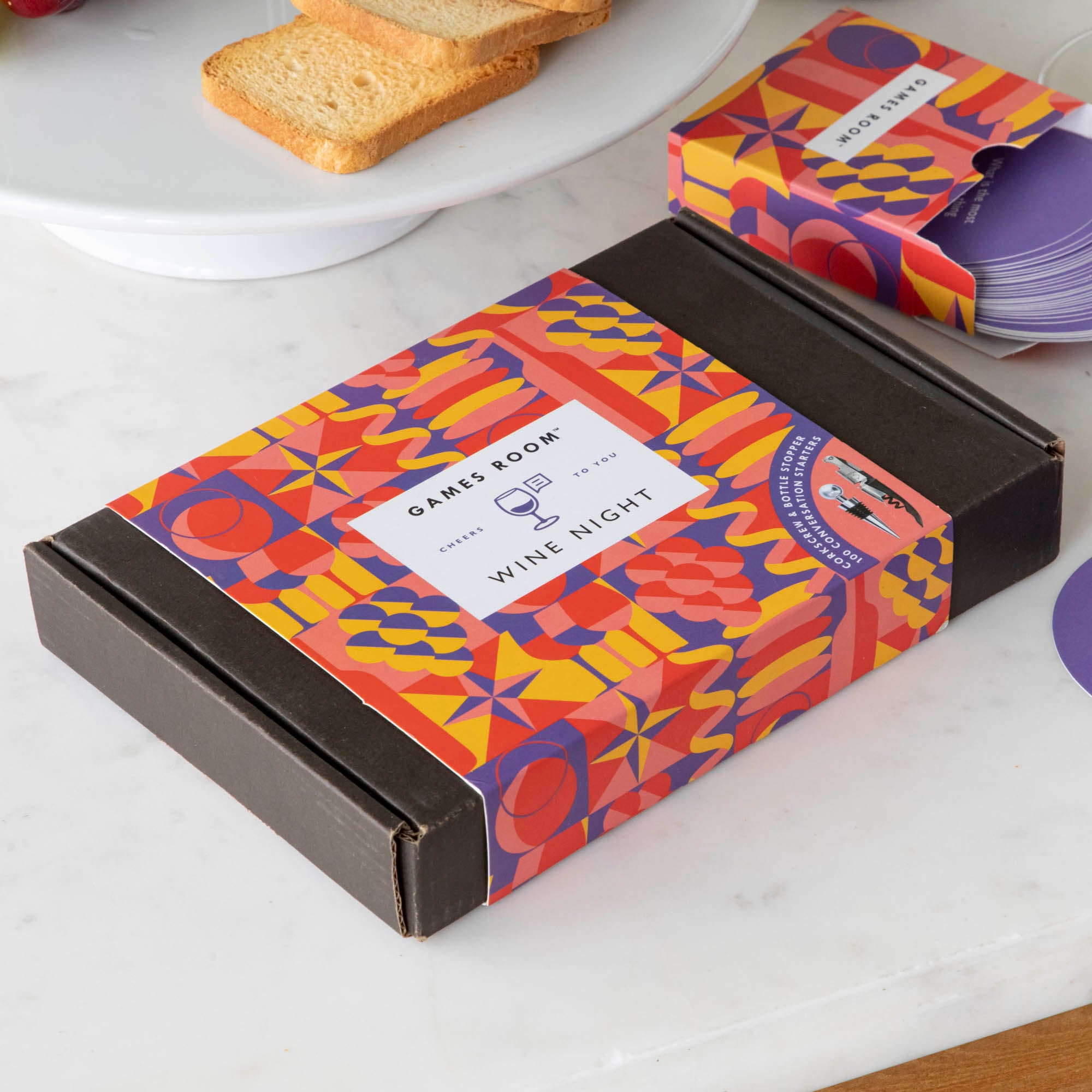 A gift set for wine nights displayed on a table, including a corkscrew, a bottle stopper, a glass of wine, a bottle of wine, bread, and grapes. The set comes in a colorful box with conversation starter cards visible.