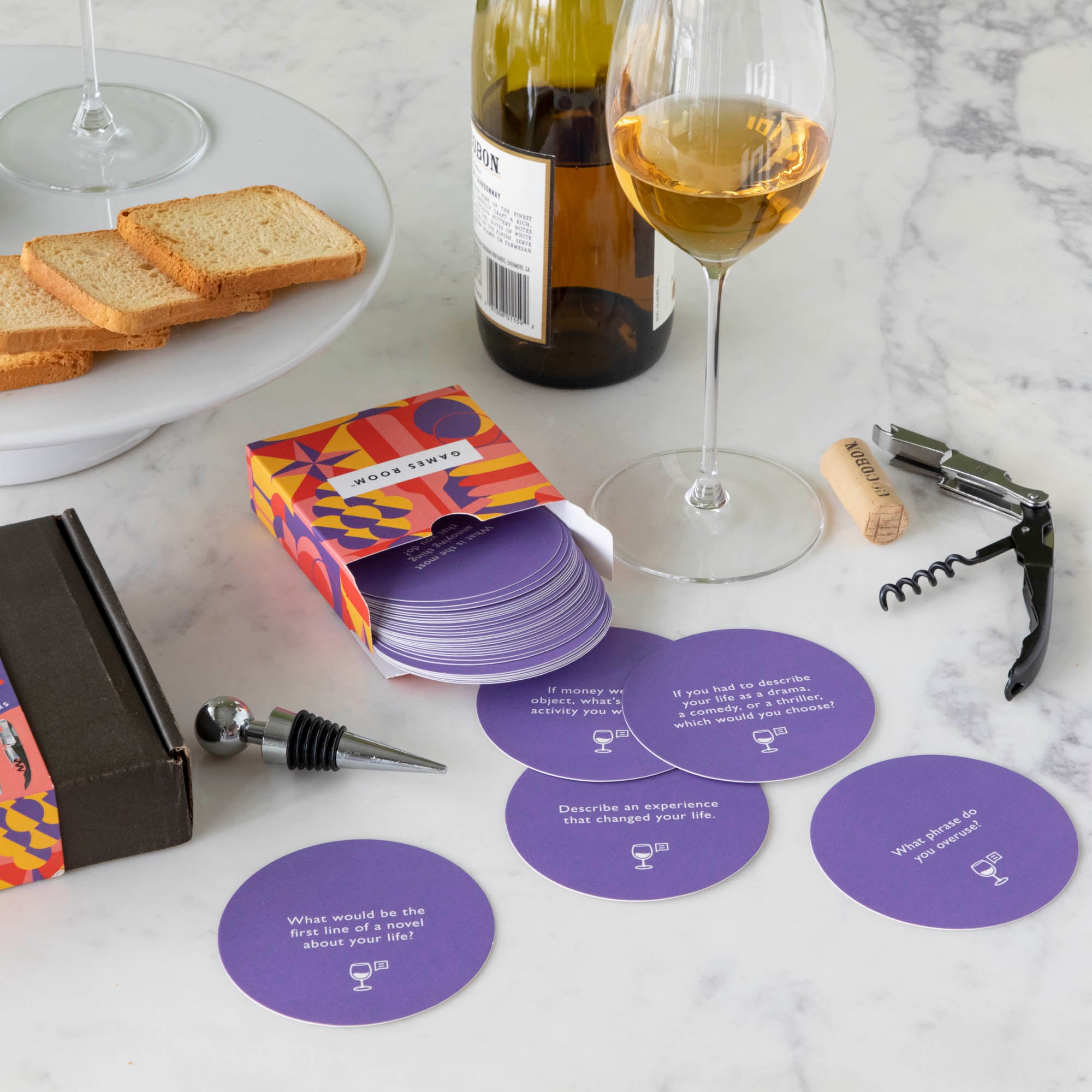 A gift set for wine nights displayed on a table, including a corkscrew, a bottle stopper, a glass of wine, a bottle of wine, bread, and grapes. The set comes in a colorful box with conversation starter cards visible.