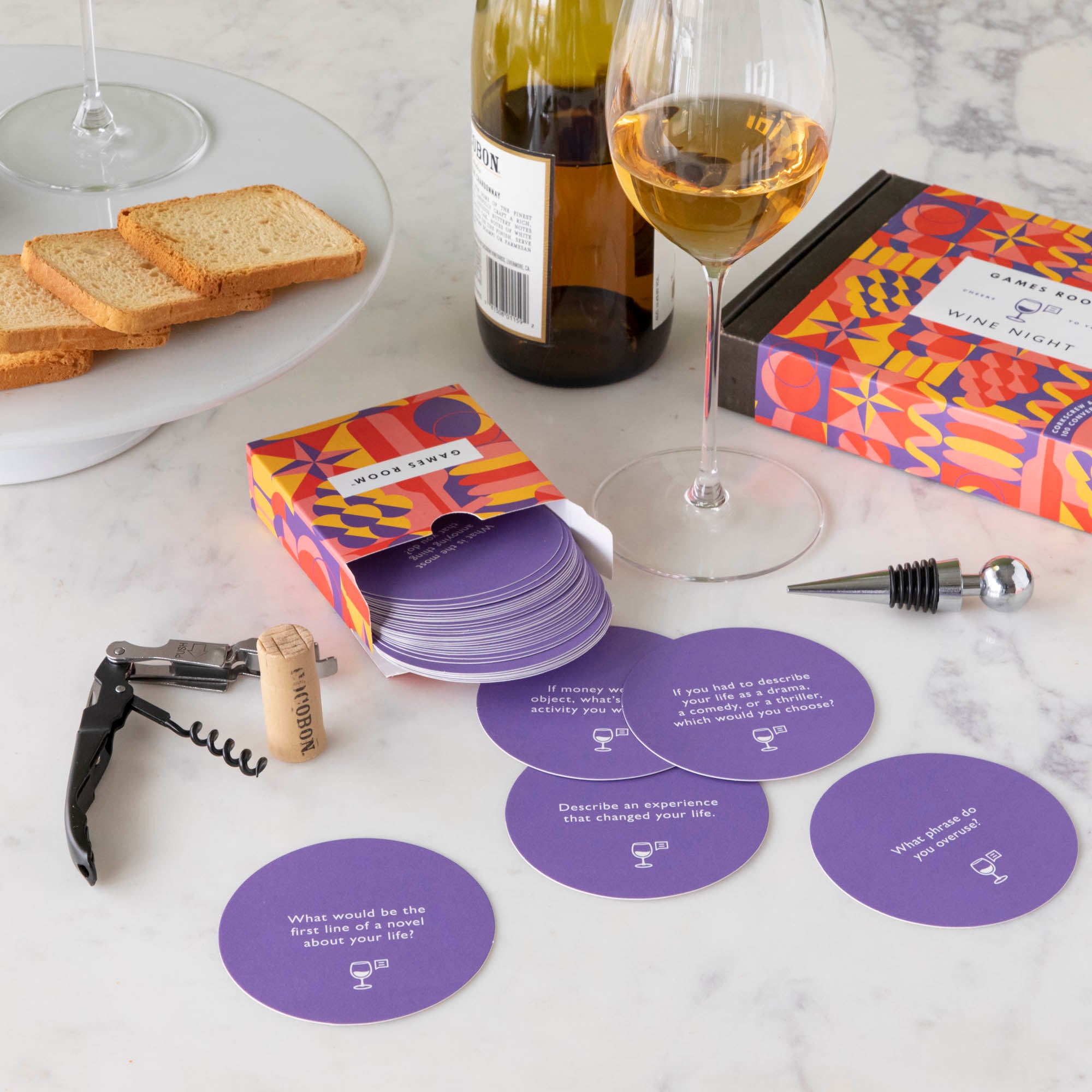 A gift set for wine nights displayed on a table, including a corkscrew, a bottle stopper, a glass of wine, a bottle of wine, bread, and grapes. The set comes in a colorful box with conversation starter cards visible.