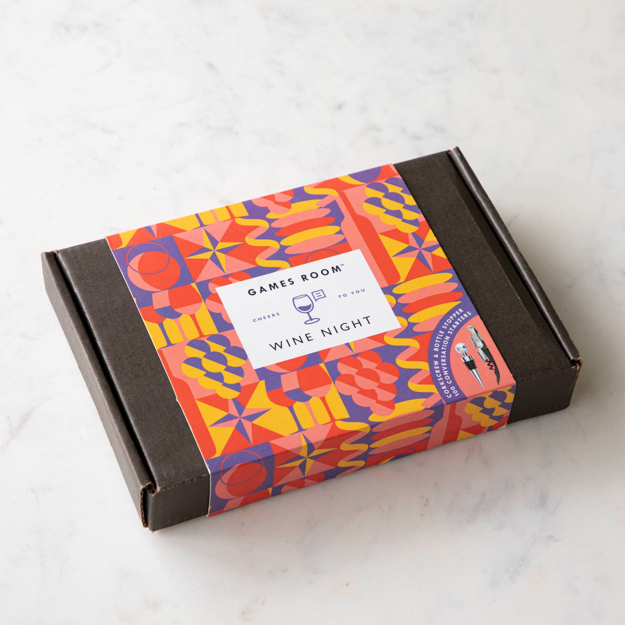 A gift set for wine nights displayed on a table, including a corkscrew, a bottle stopper, a glass of wine, a bottle of wine, bread, and grapes. The set comes in a colorful box with conversation starter cards visible.