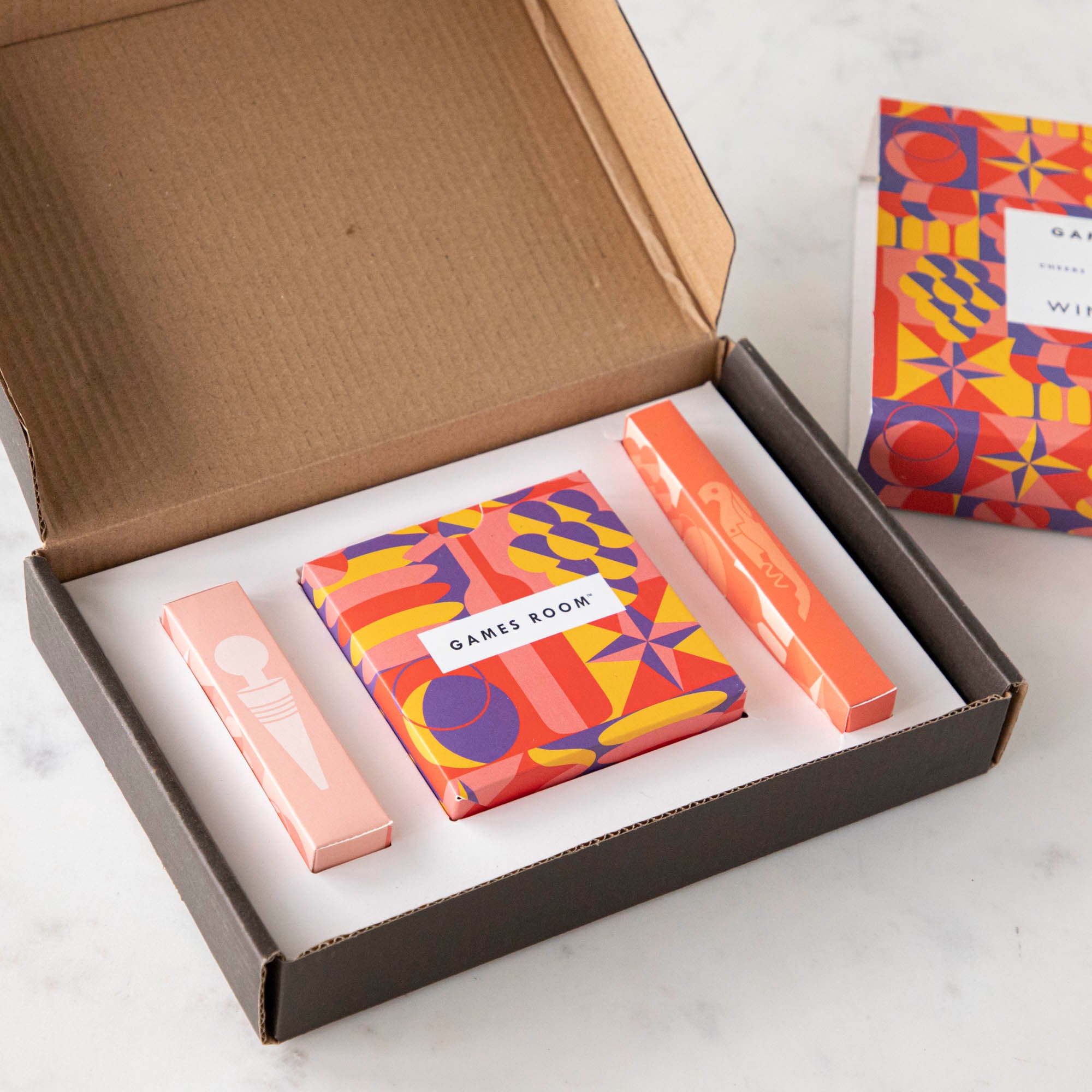 A gift set for wine nights displayed on a table, including a corkscrew, a bottle stopper, a glass of wine, a bottle of wine, bread, and grapes. The set comes in a colorful box with conversation starter cards visible.