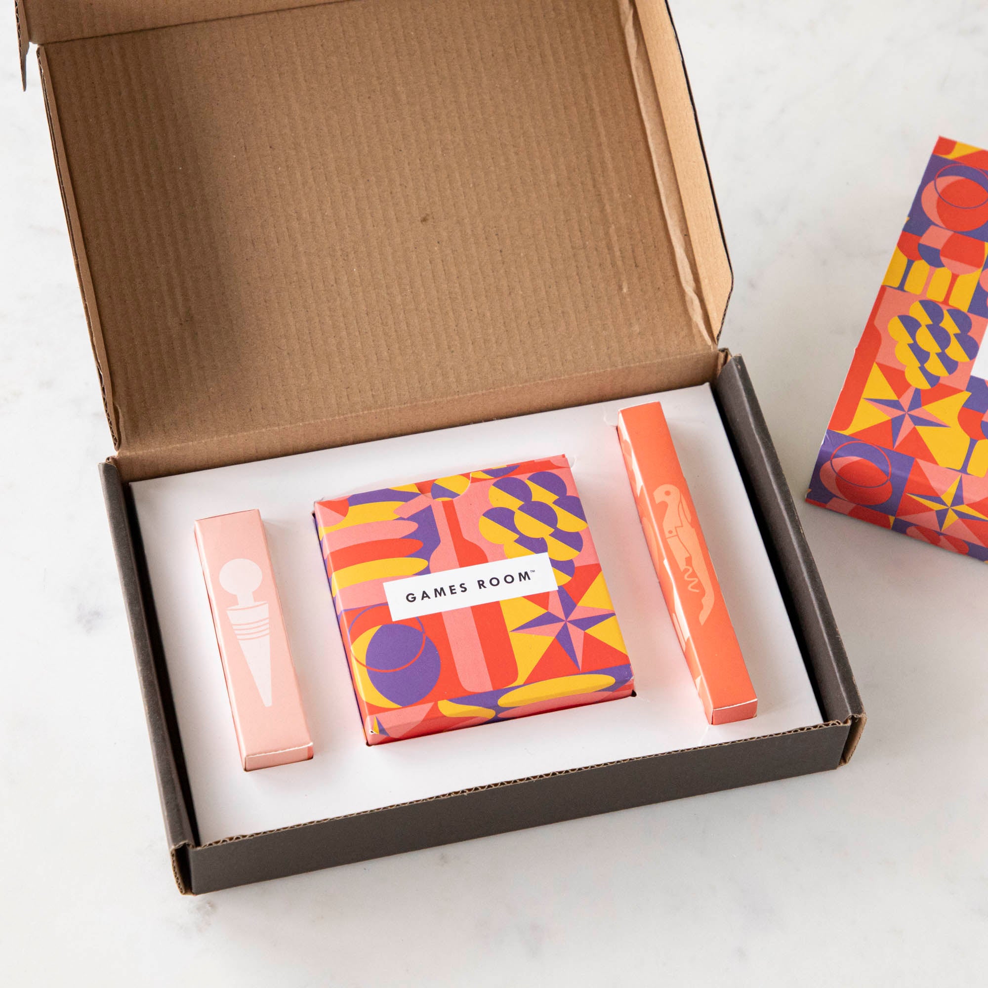 A gift set for wine nights displayed on a table, including a corkscrew, a bottle stopper, a glass of wine, a bottle of wine, bread, and grapes. The set comes in a colorful box with conversation starter cards visible.