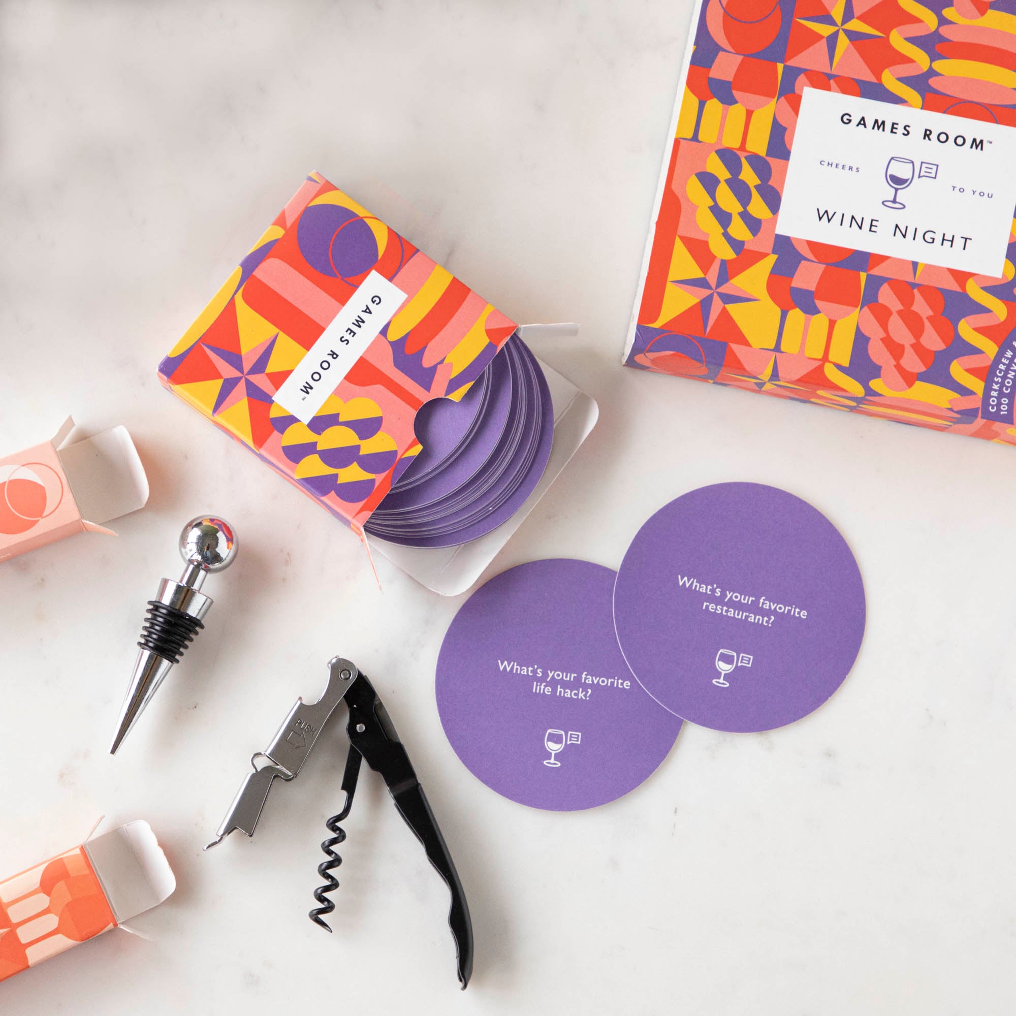 A gift set for wine nights displayed on a table, including a corkscrew, a bottle stopper, a glass of wine, a bottle of wine, bread, and grapes. The set comes in a colorful box with conversation starter cards visible.