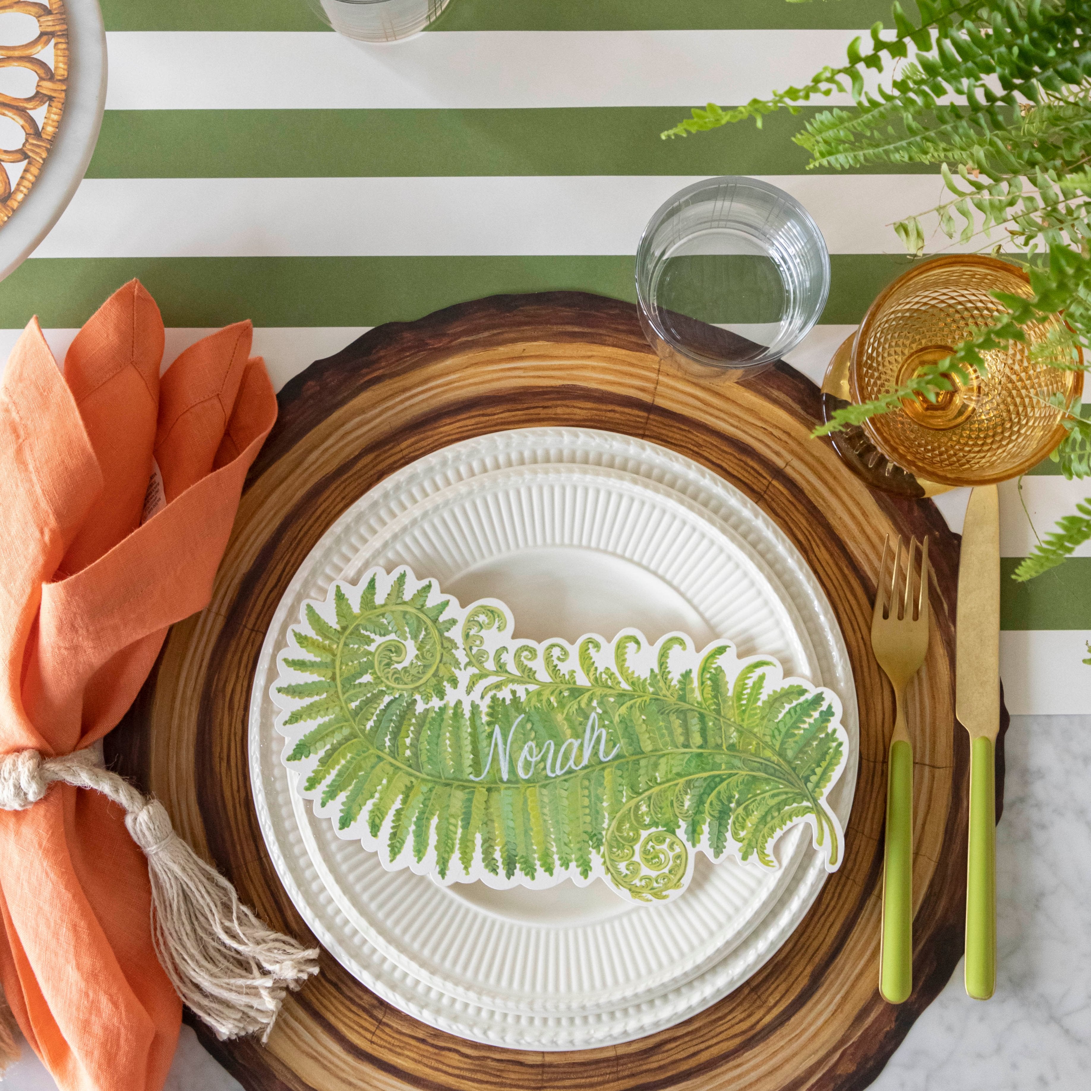 Table setting with a Die-cut Wood Slice Placemat on a green stripe runner, with white plates and a Fern table accent with the name "Noah" written on it on top.