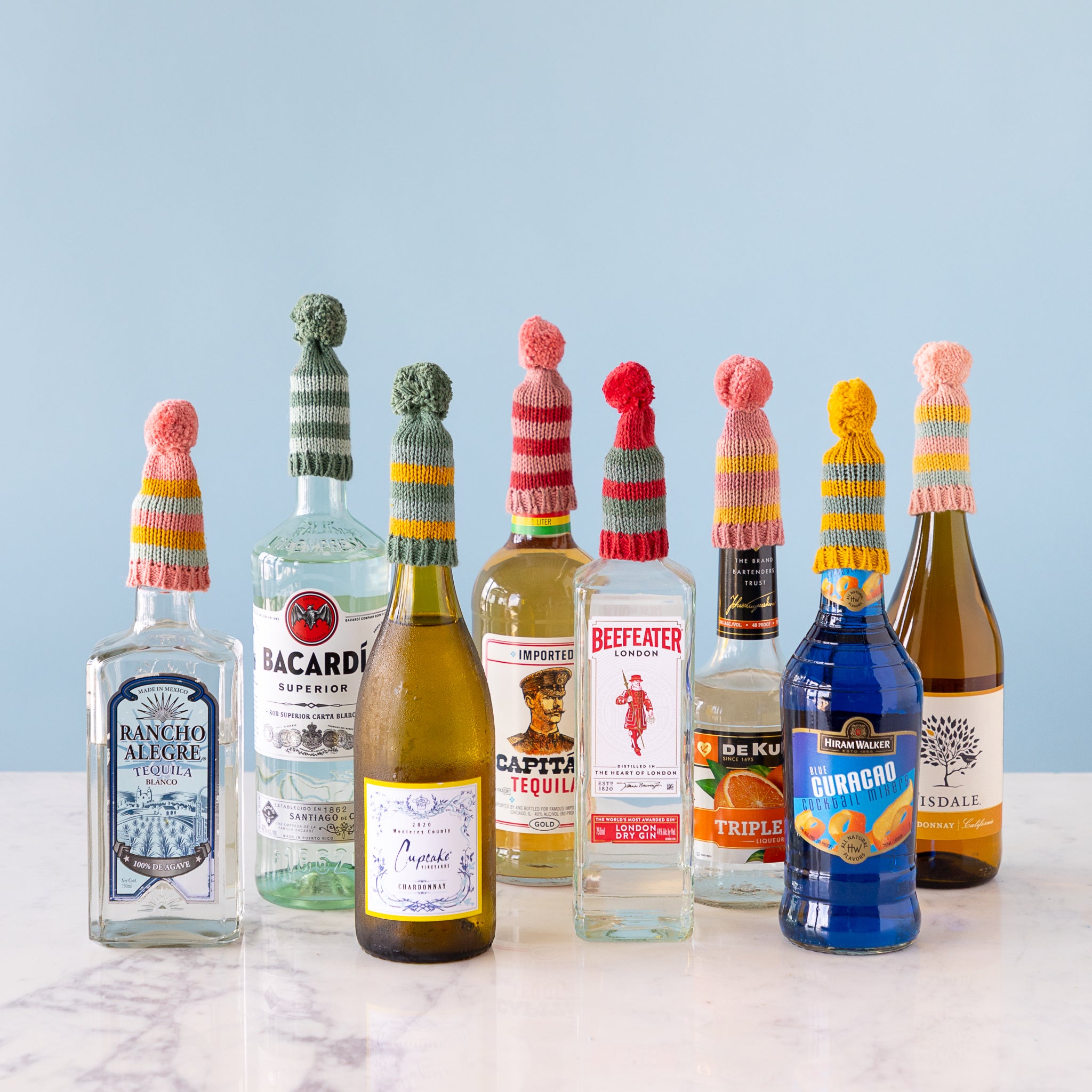 Bottles of alcohol with colorful knit hats on a light blue background.