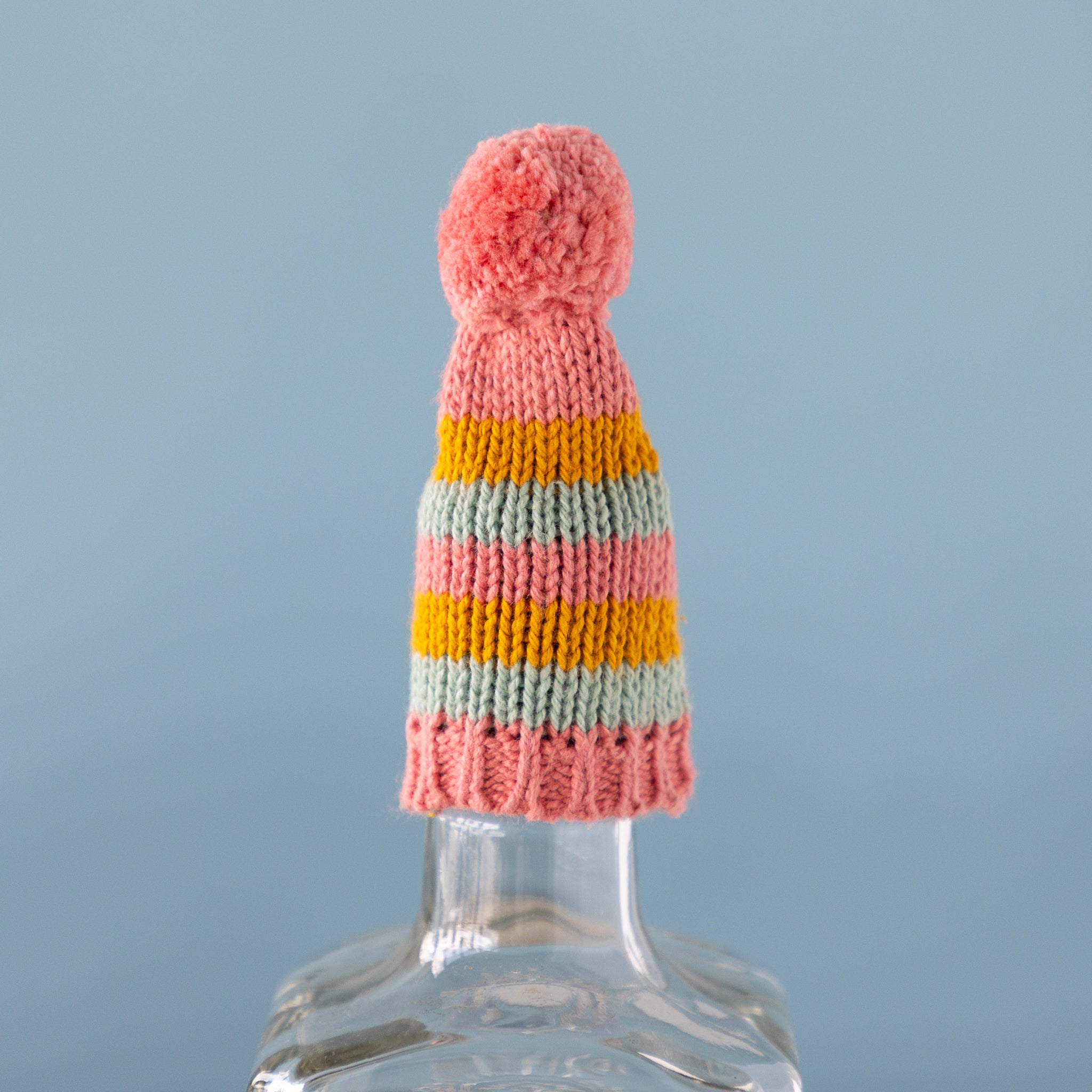 Colorful knitted hat with a pom-pom on a clear bottle against a light blue background.