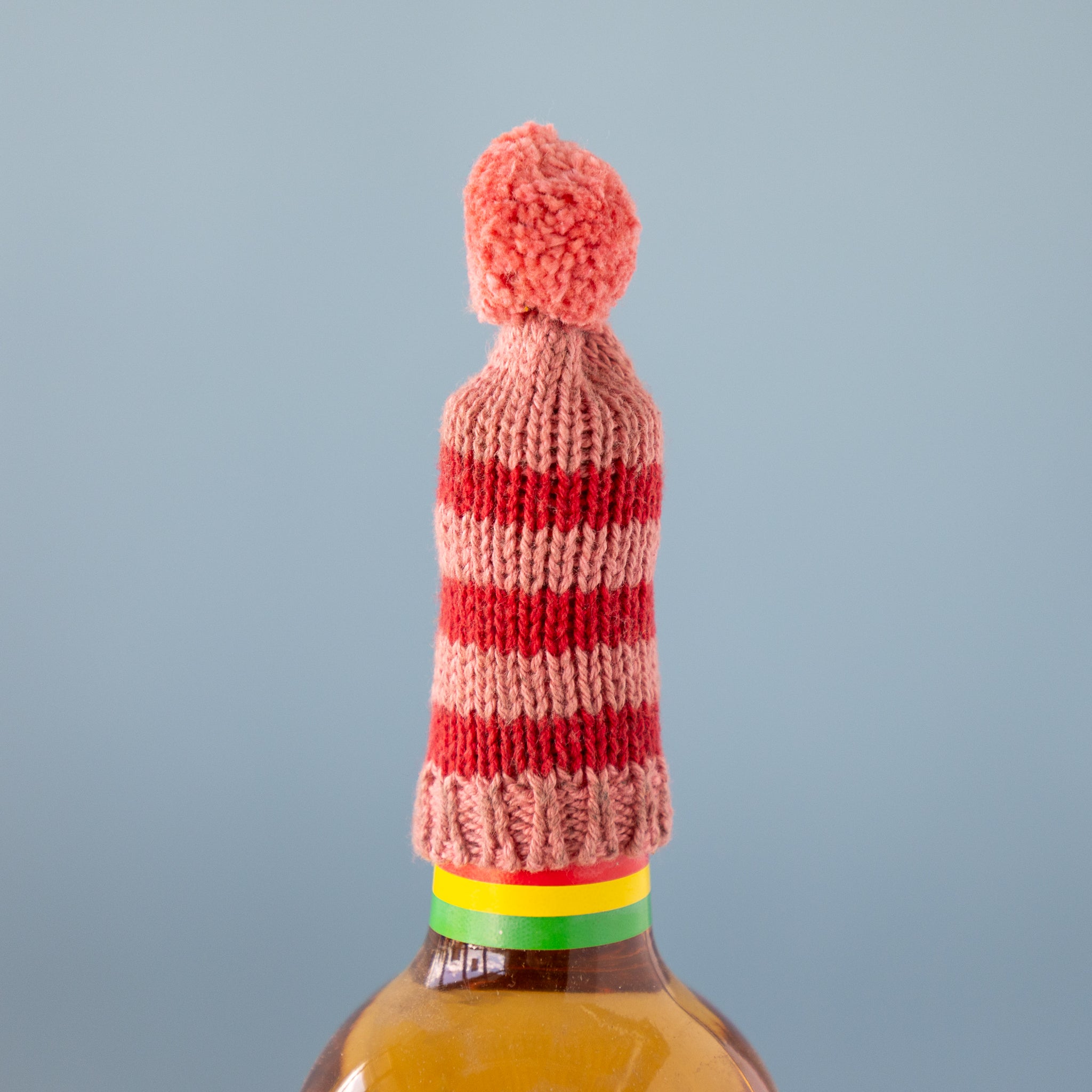 Knitted bottle cover with a red and pink striped pattern and a pom-pom on a bottle against a gray background.