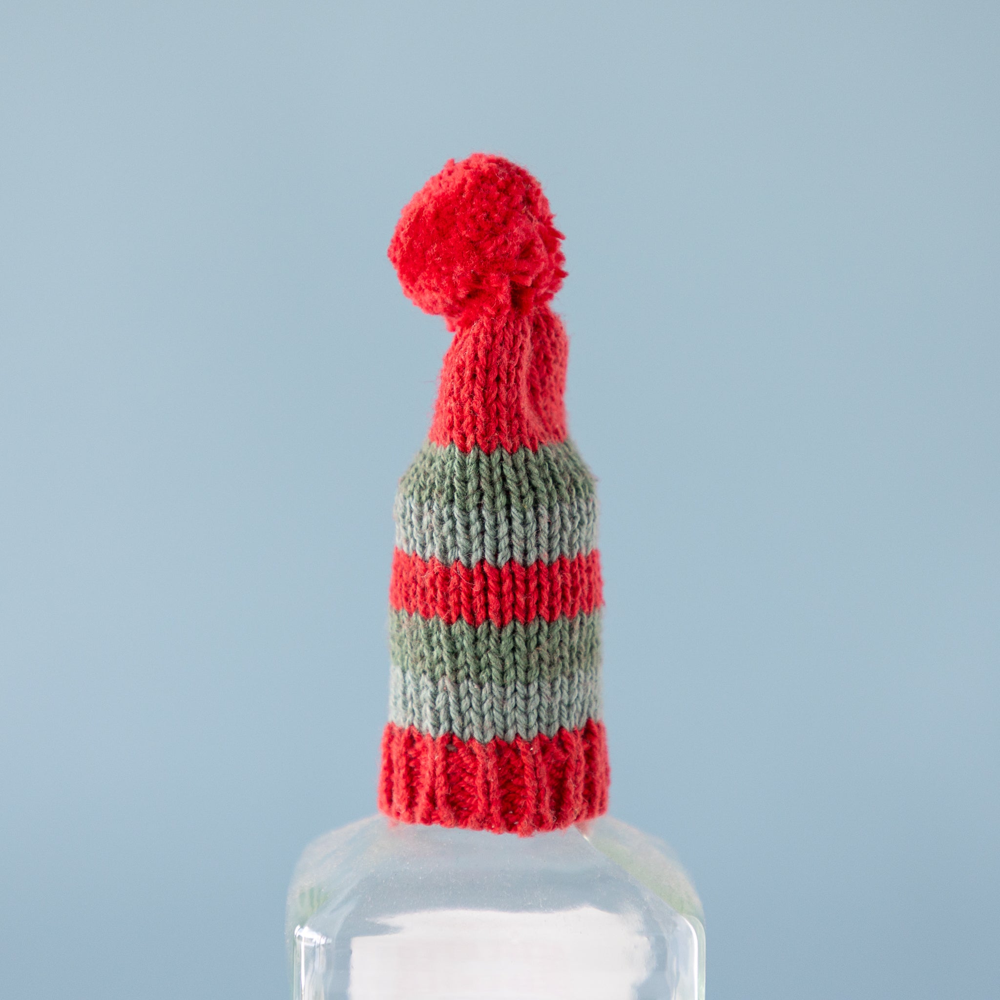 Knitted hat with red and green pom-pom and stripes on a clear bottle against a light blue background.