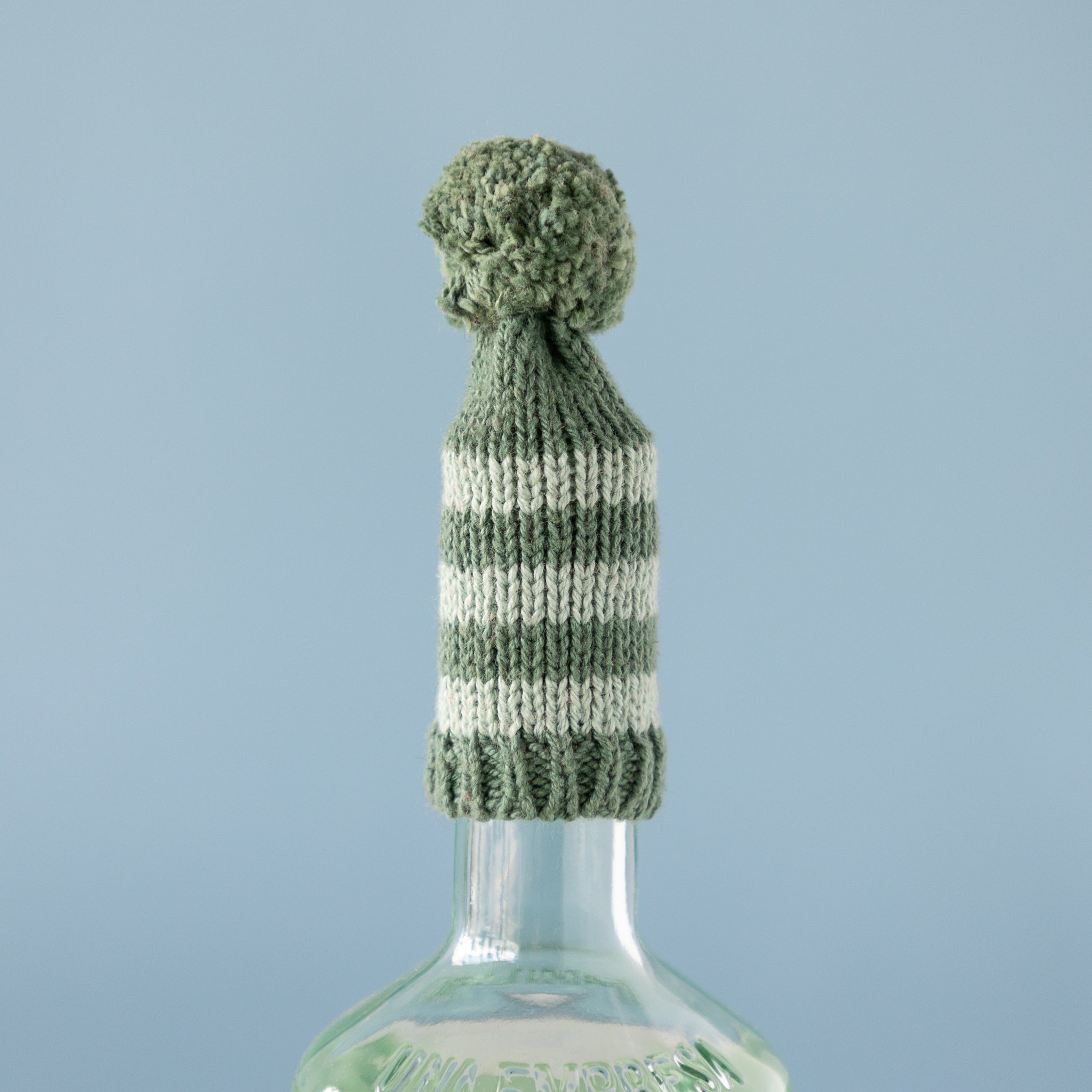 Knitted bottle cover with a pom-pom on a clear bottle against a light blue background.