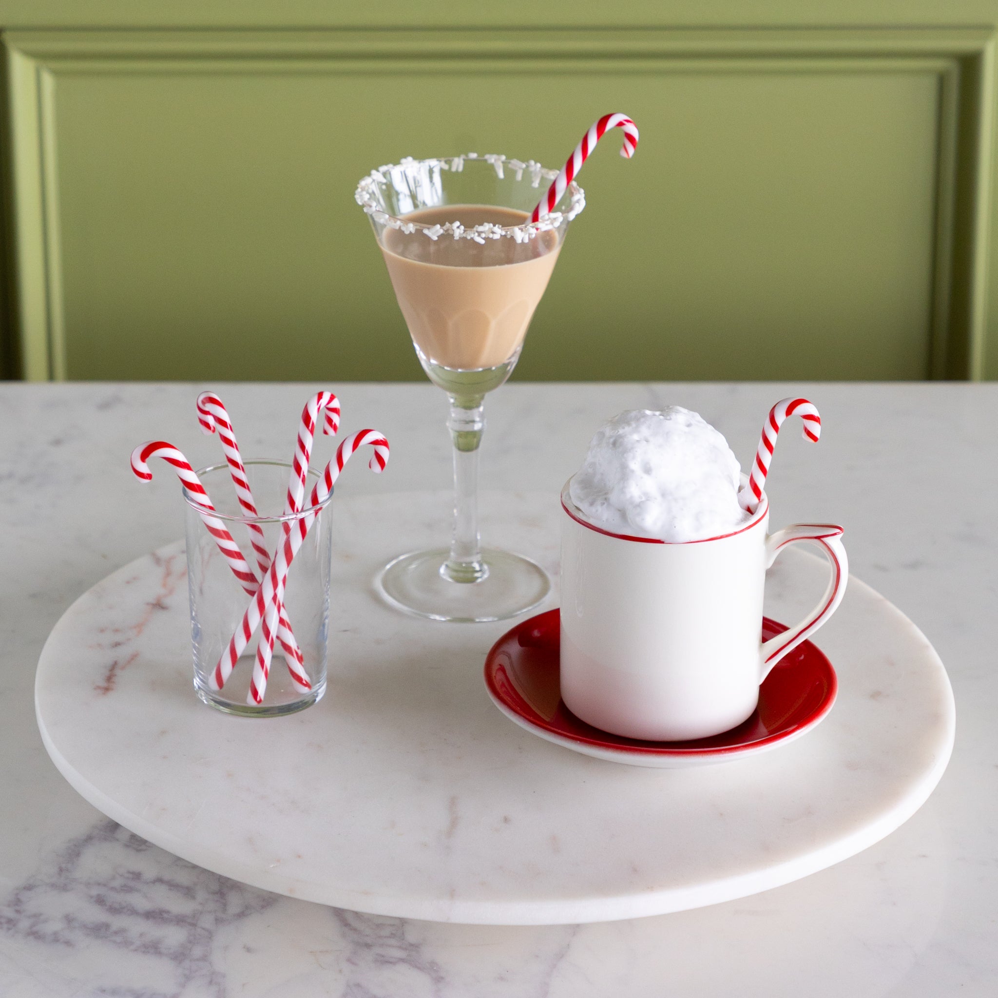 A mug with whipped cream, a mocha cocktail and a small glass with candy cane stir sticks in them on a marble lazy susan.
