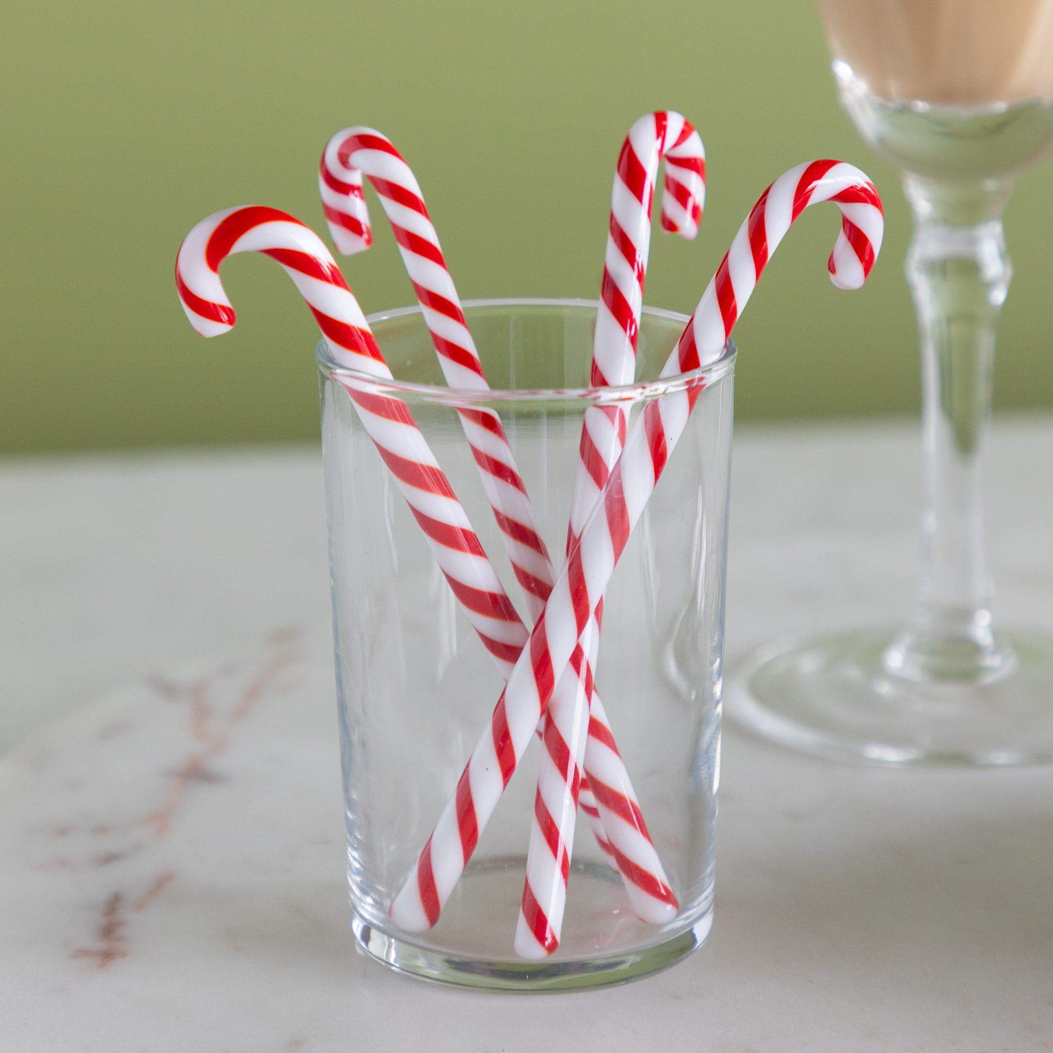 Red and white candy cane stir sticks in a clear glass on a marble surface with a green background.