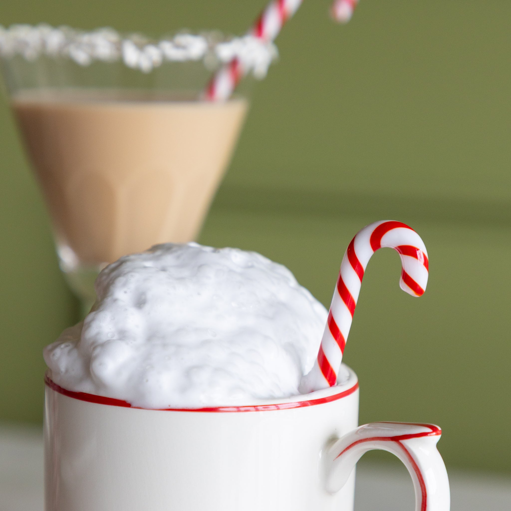 Mug with whipped cream and a candy cane stir stick on a green background.