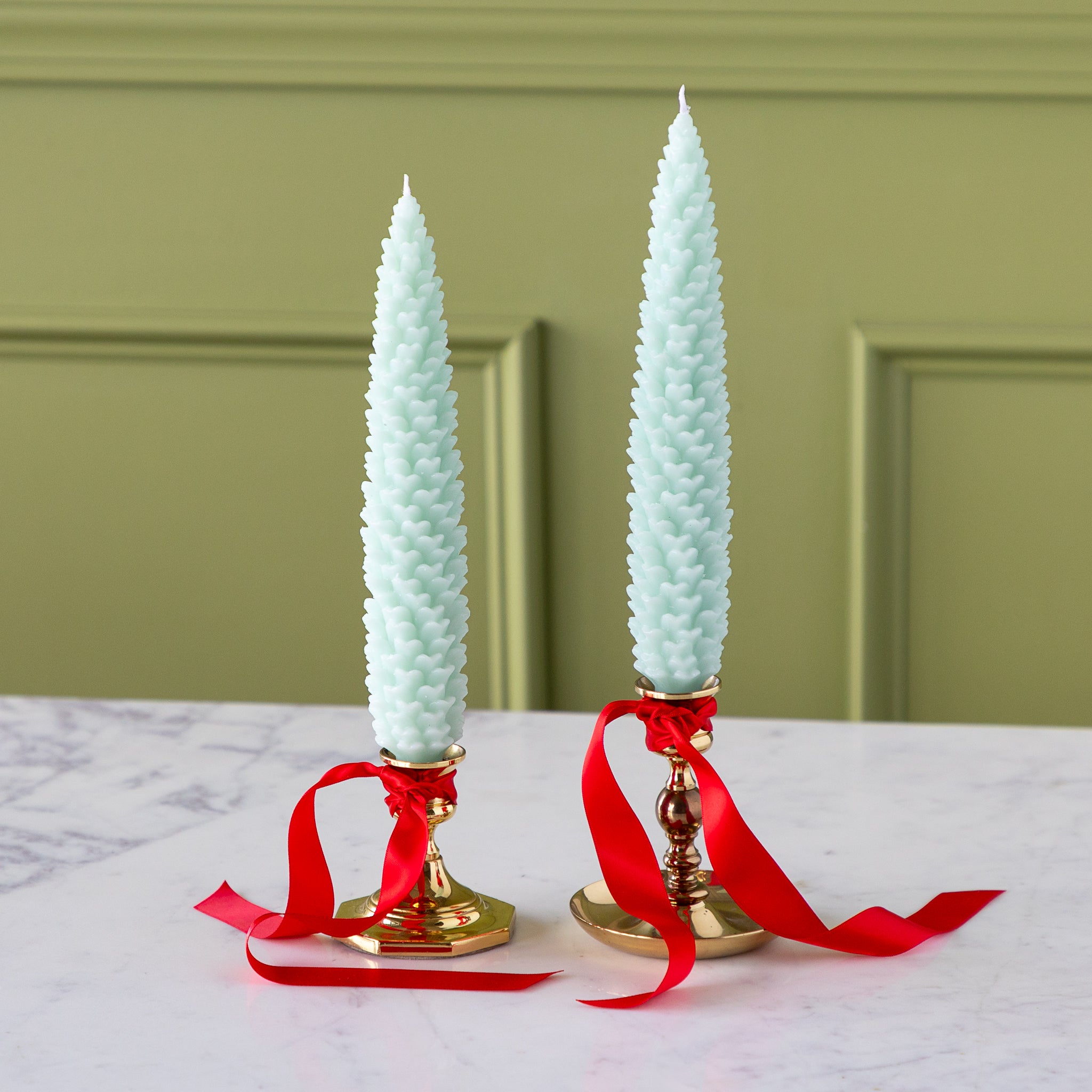 Two Mint Large Pinecone Taper Candles in gold candlesticks with red ribbons on a marble surface.