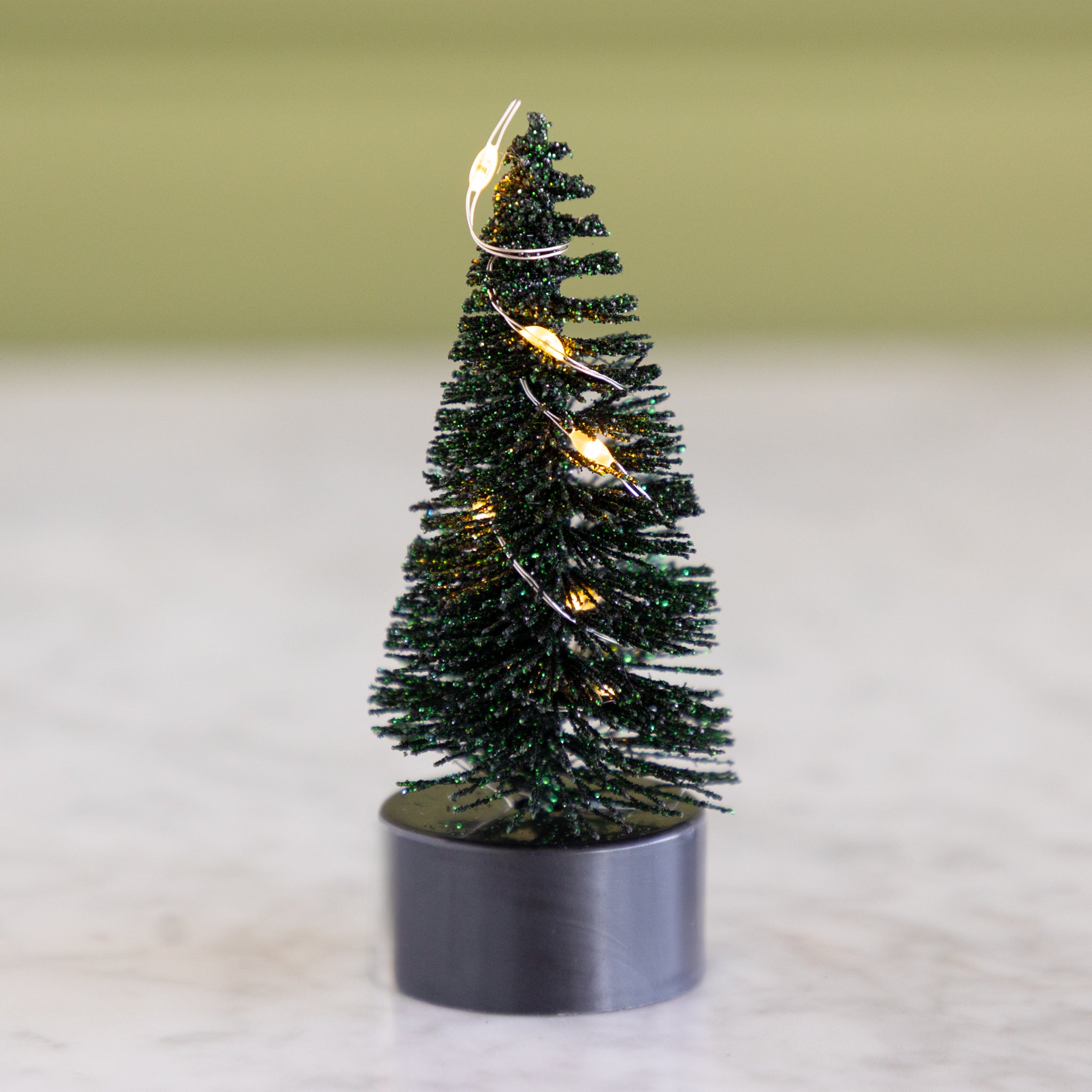 Small forest green bottle-brush tree with lights on a neutral background.