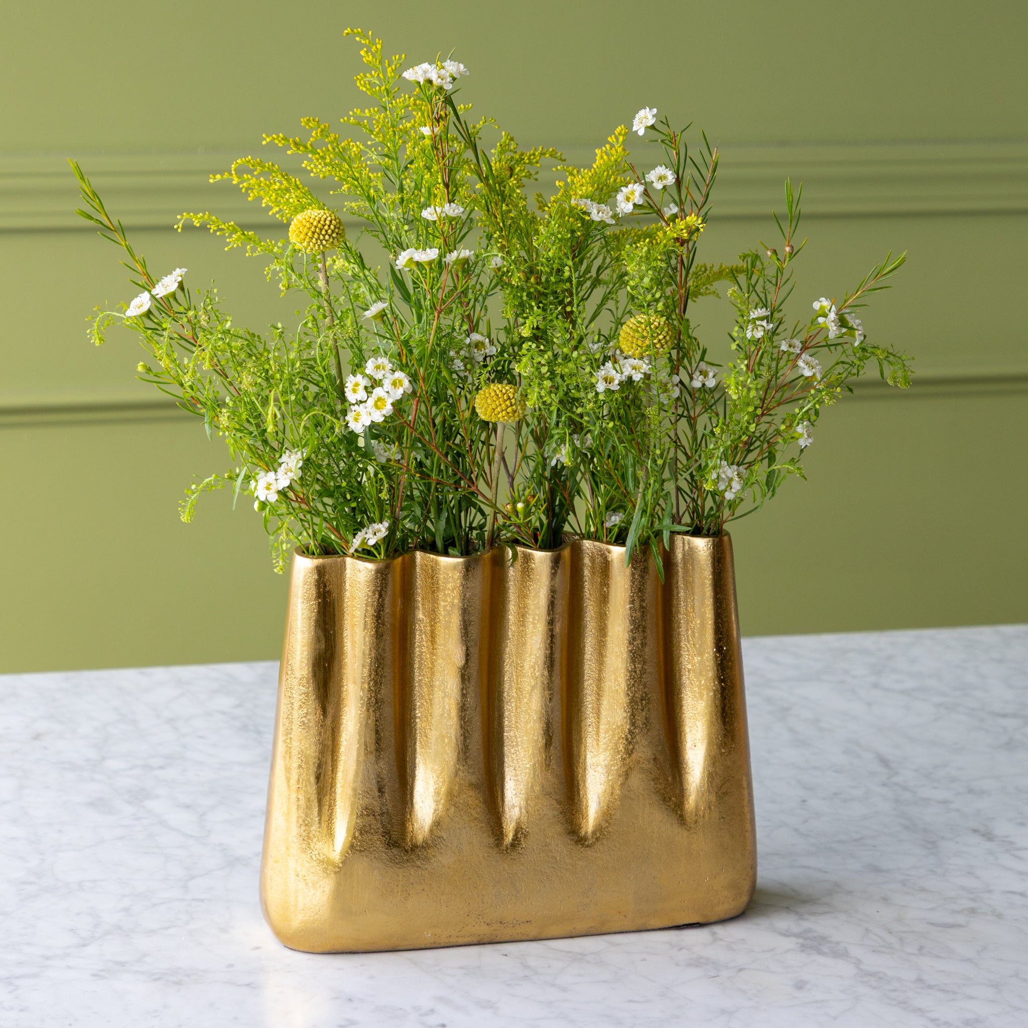 Tall Aluminum Gold Wave vase with greenery on a light surface against a green wall.