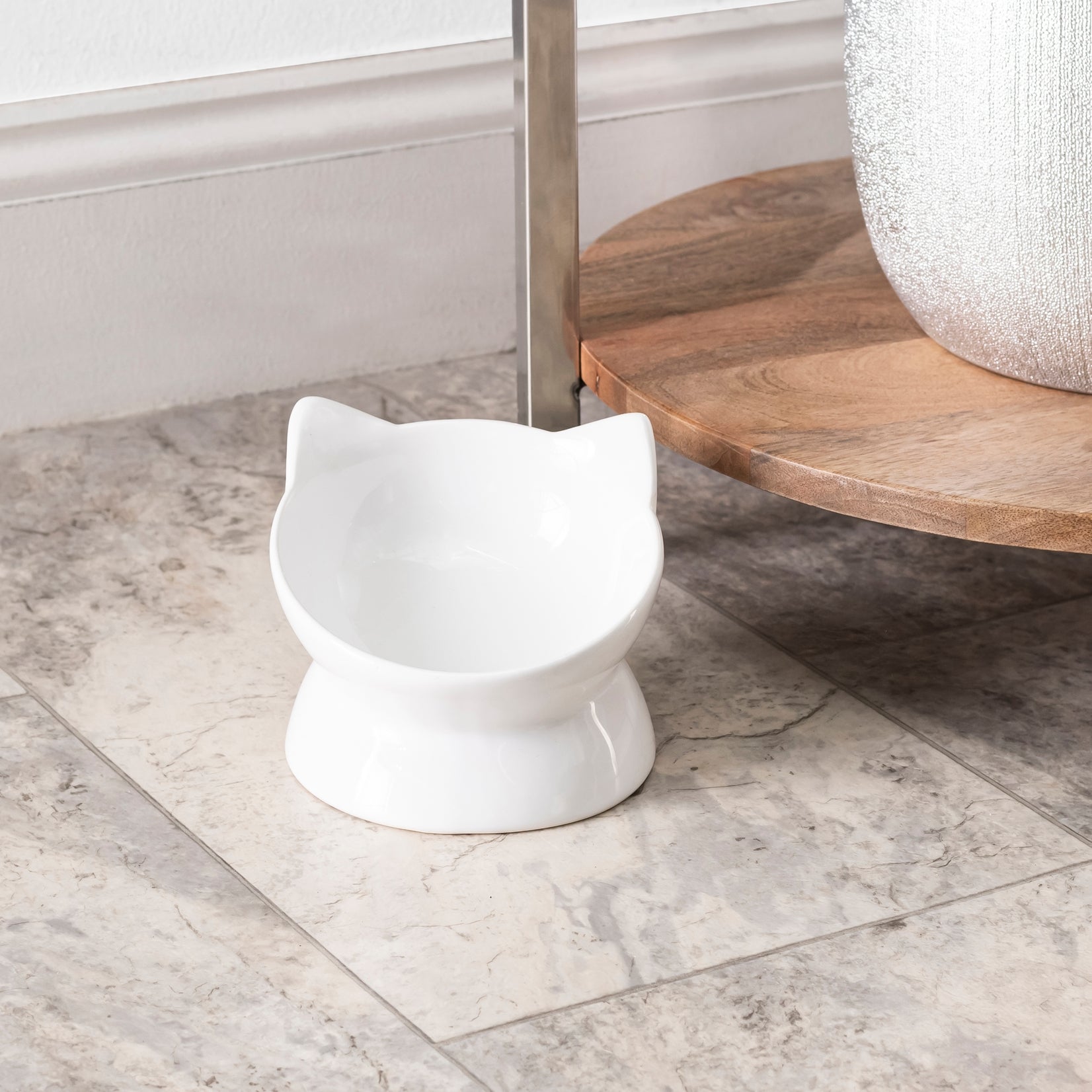 White cat-shaped food bowl on a marble floor with a wooden shelf in the background.