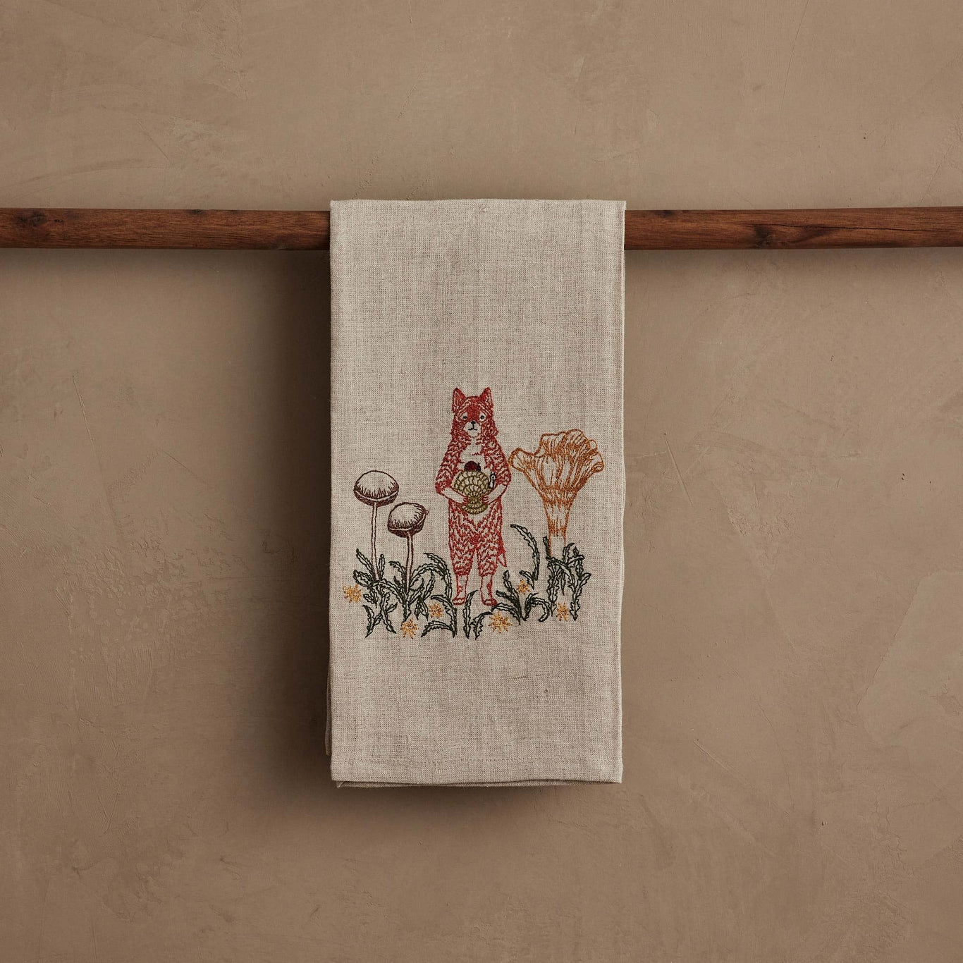 Tea towel with fox and mushroom design hanging on a wooden rod against a beige wall.