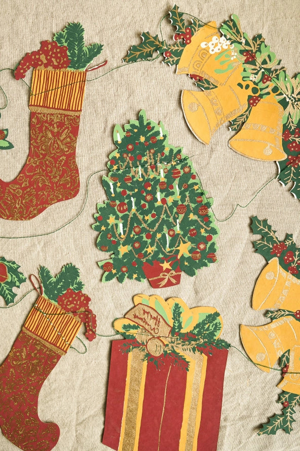 A close-up of a Christmas-themed paper garland print featuring red stockings, a decorated green Christmas tree, yellow bells, holly leaves, and a red-and-gold gift box on a light beige background.