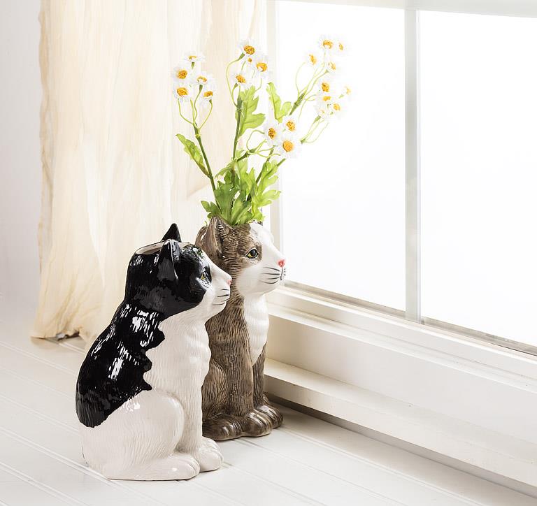 Two cat shaped ceramic vases, one black and white one brown and white, sitting on a surface next to a window as if they are gazing outside.