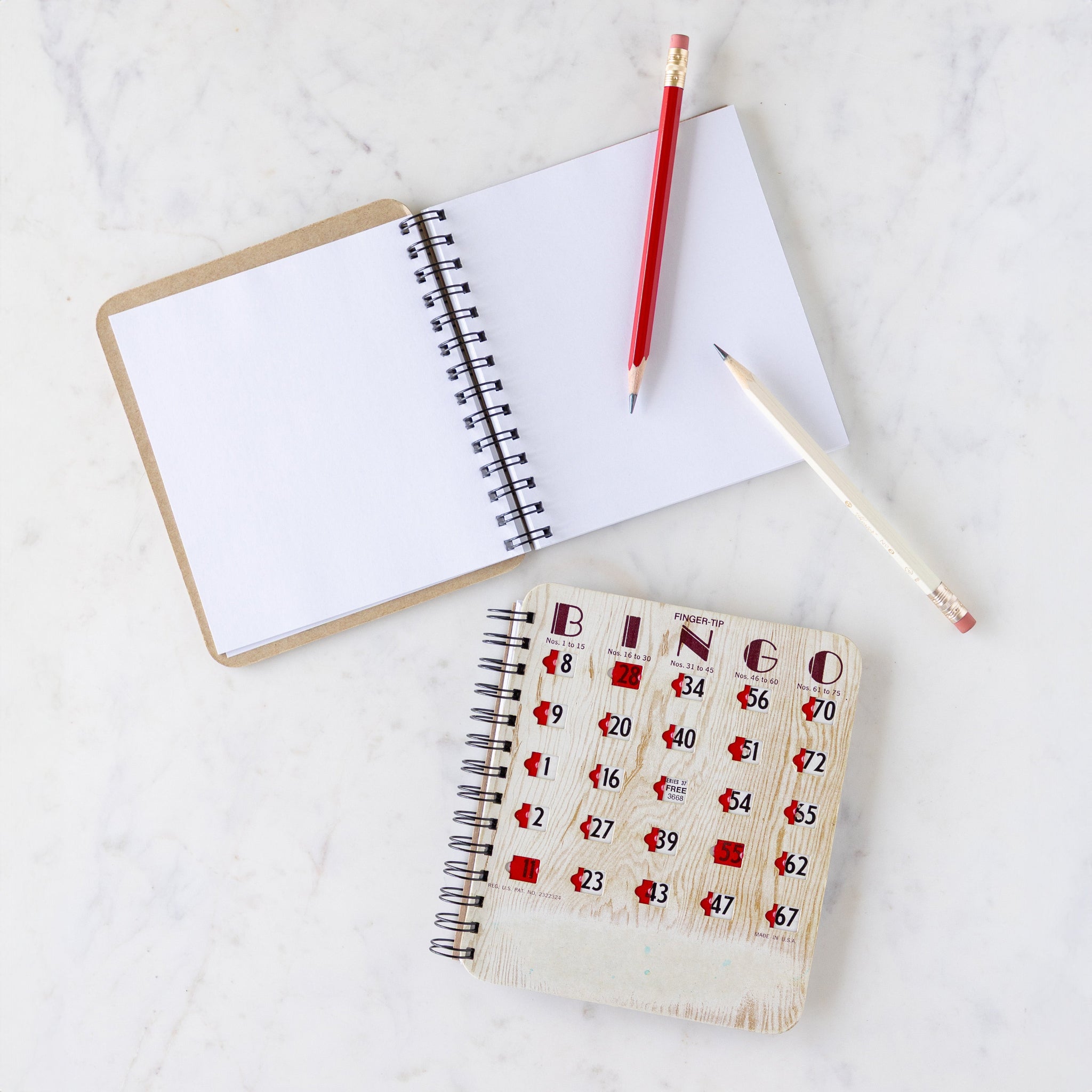 Open Bingo Notebook with two pencils atop, next to a closed Bingo Book featuring a classic bingo board cover with red markers to cover the numbers.