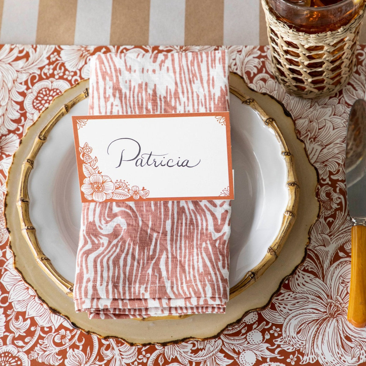 A folded woodgrain-patterned linen napkin in a coral/pink and white color laying atop a white ceramic plate with a matching place card placed atop it.