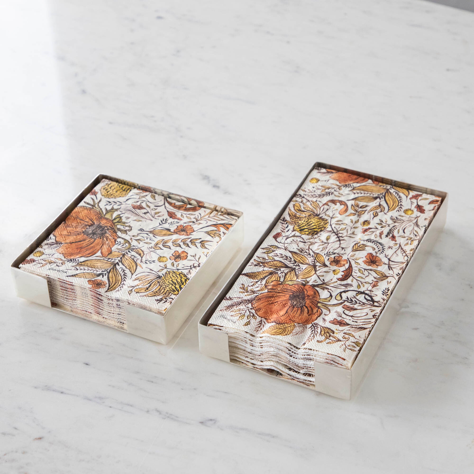 Two Silver Napkin Holders, guest-sized and cocktail-sized, containing autumnal napkins on a white table.