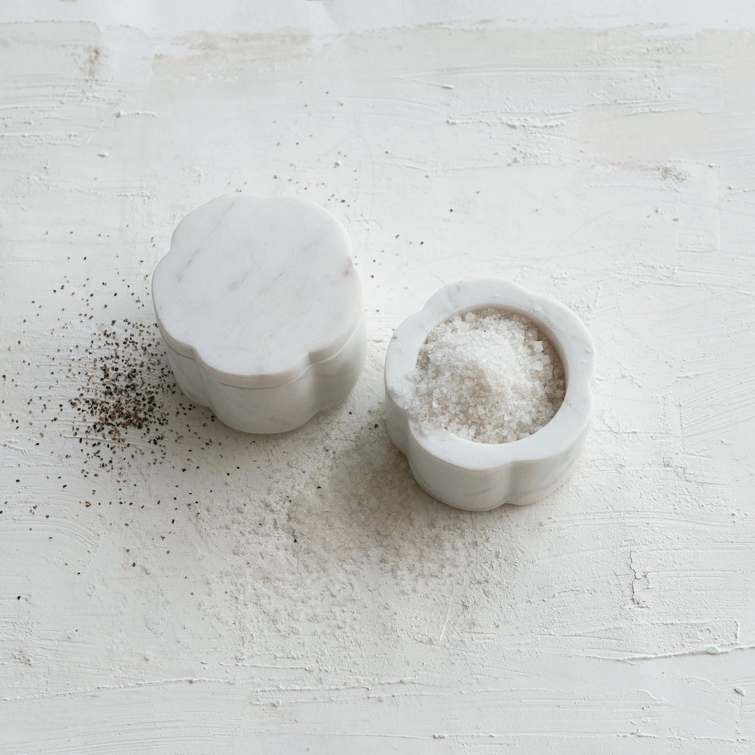 Two scalloped marble containers on a white surface one with a lid on and one open and full of salt.