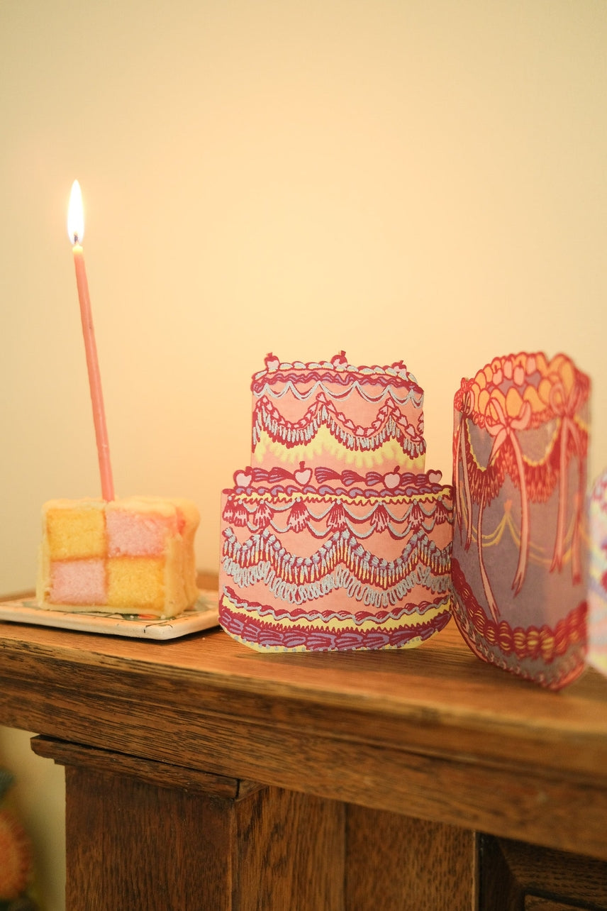 Cake greeting card with pink candle and decorative boxes on a wooden surface.