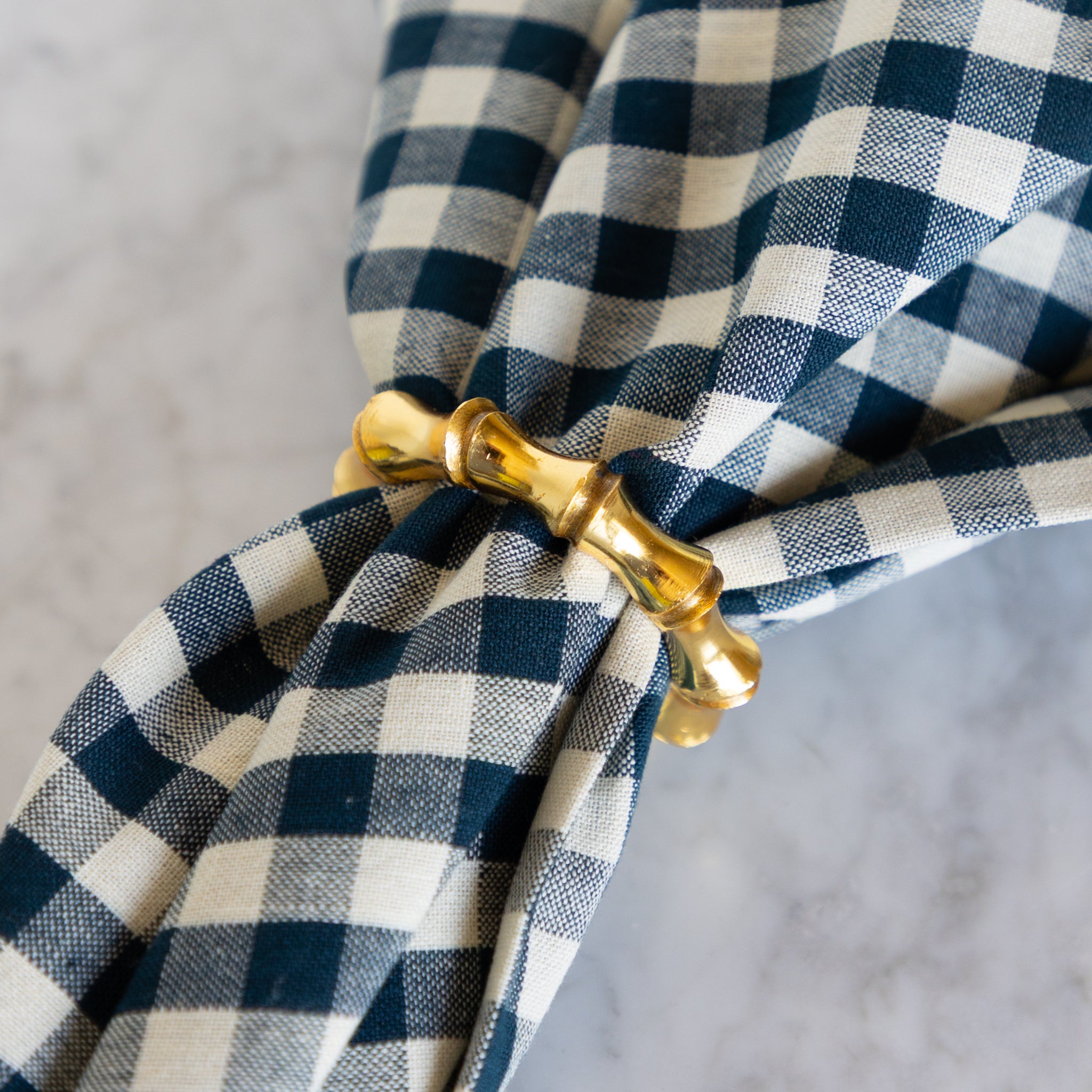 Blue and white checkered napkin with a gold napkin ring on a gray background.