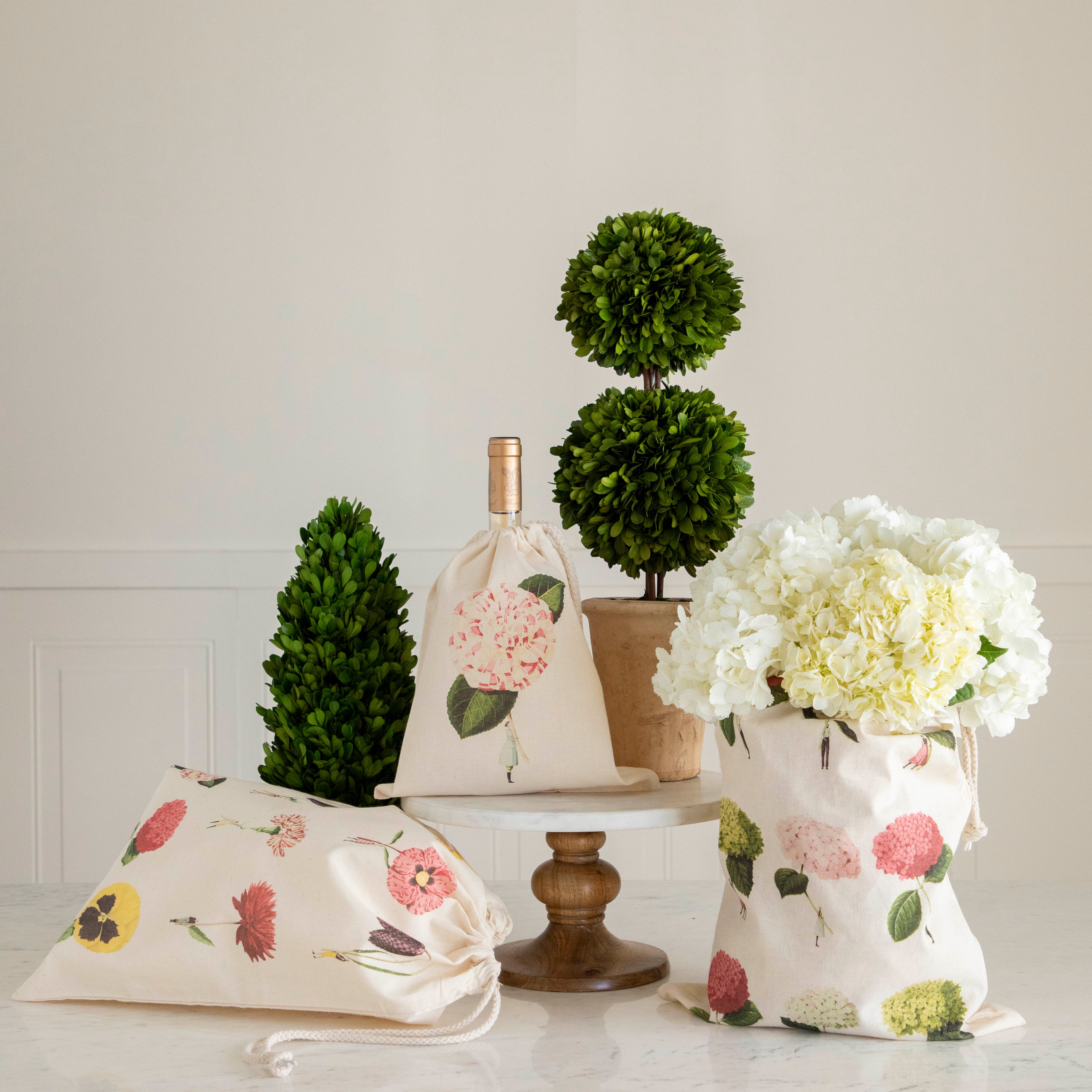 Decorative indoor setting with Laura Stoddart drawstring bags in floral patterns, topiary plants, and a vase of white hydrangeas on a table from Hester &amp; Cook.
