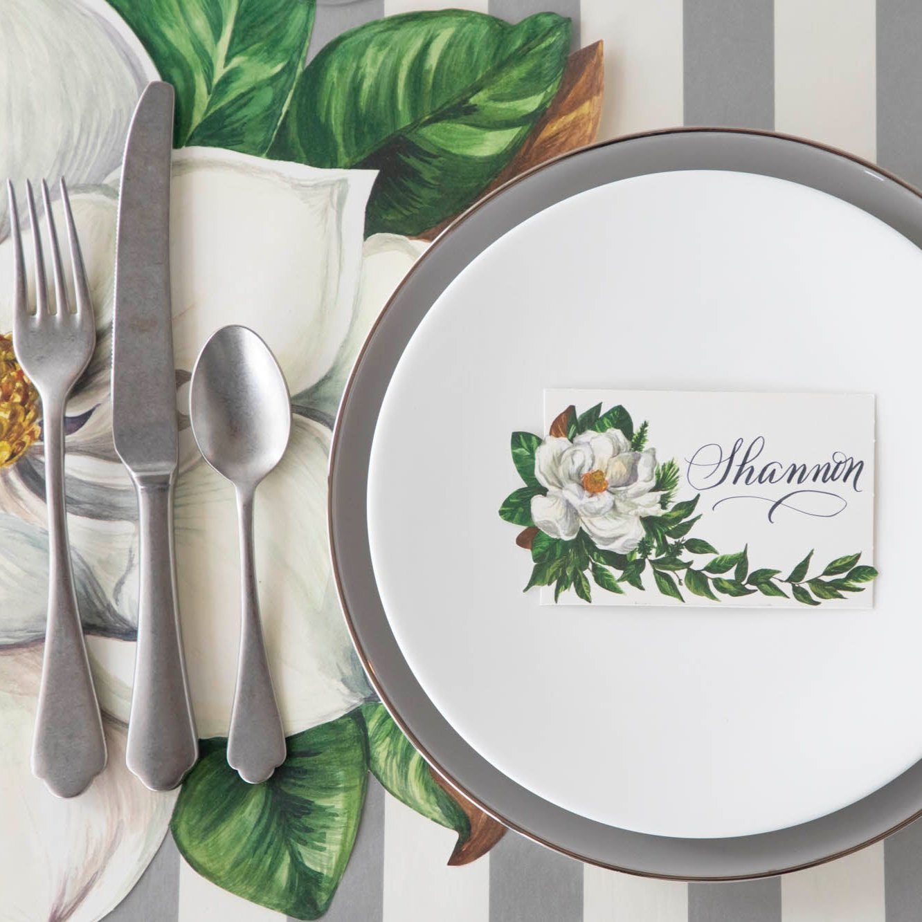 An elegant table setting with the Magnolia Place Card with "Shannon" written on it, on a set of dinner plates, next to the Die-cut Magnolia Placemat and Silver Classic Stripe Runner underneath.