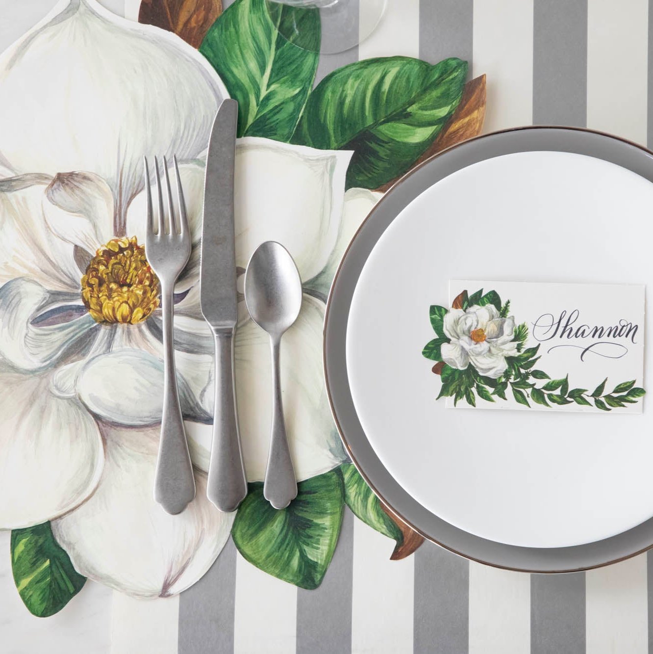 Die-cut Magnolia Placemat with silverware on top, next to a table setting with a Magnolia Place Card with the name "Shannon" written on it.