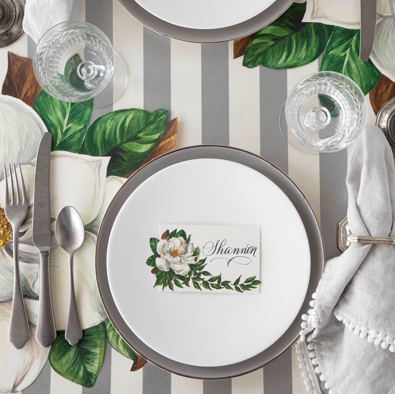 An elegant table setting with the Magnolia Place Card with "Shannon" written on it, on a set of dinner plates, next to the Die-cut Magnolia Placemat and Silver Classic Stripe Runner underneath.