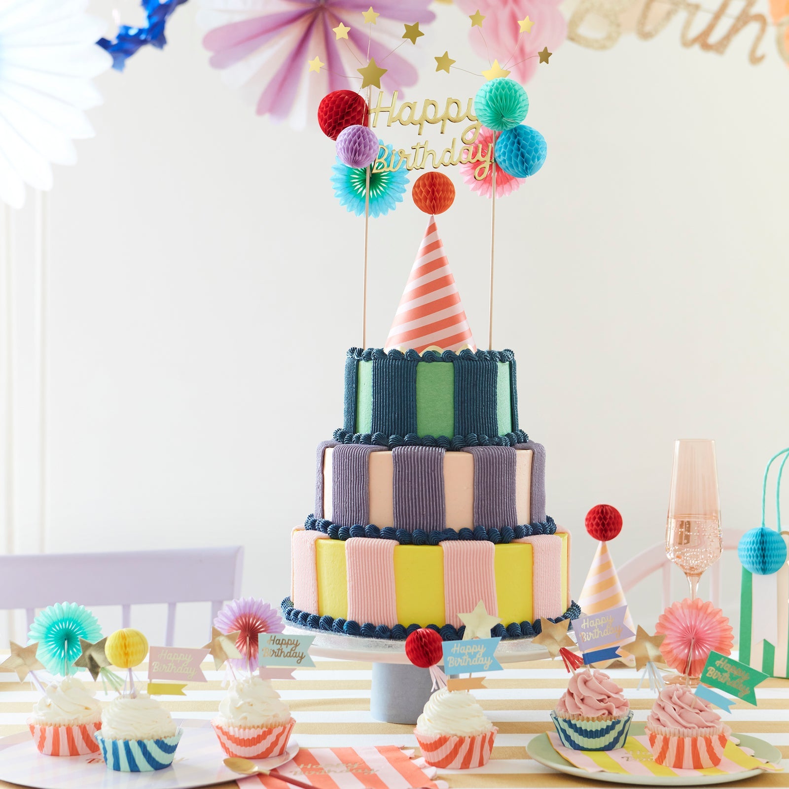 A colorful, birthday table setting with a pastel striped cake adorned with the Happy Birthday Honeycomb Cake Topper and party hat.