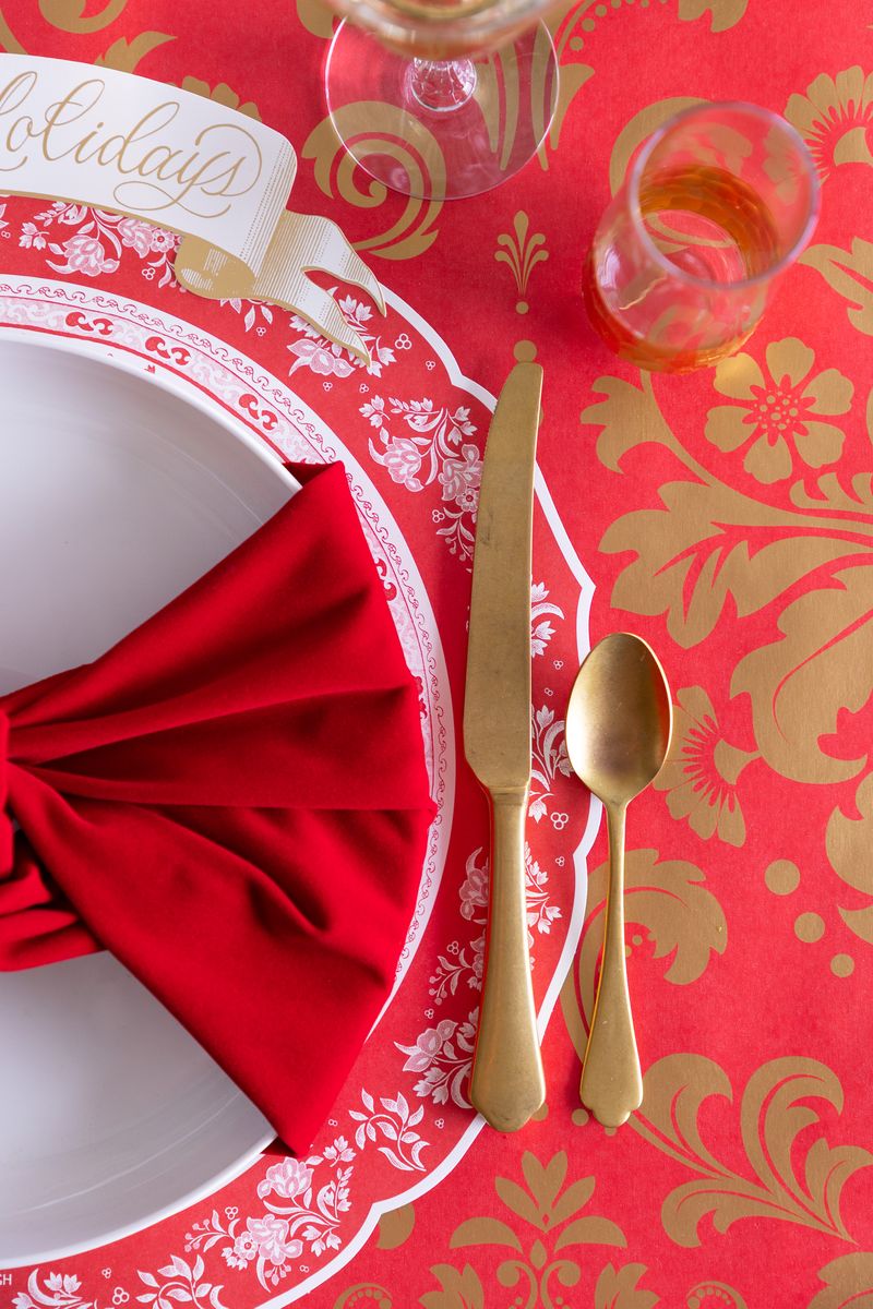 Christmas table setting with red napkin, gold cutlery, and white plate on a red and gold patterned table runner.